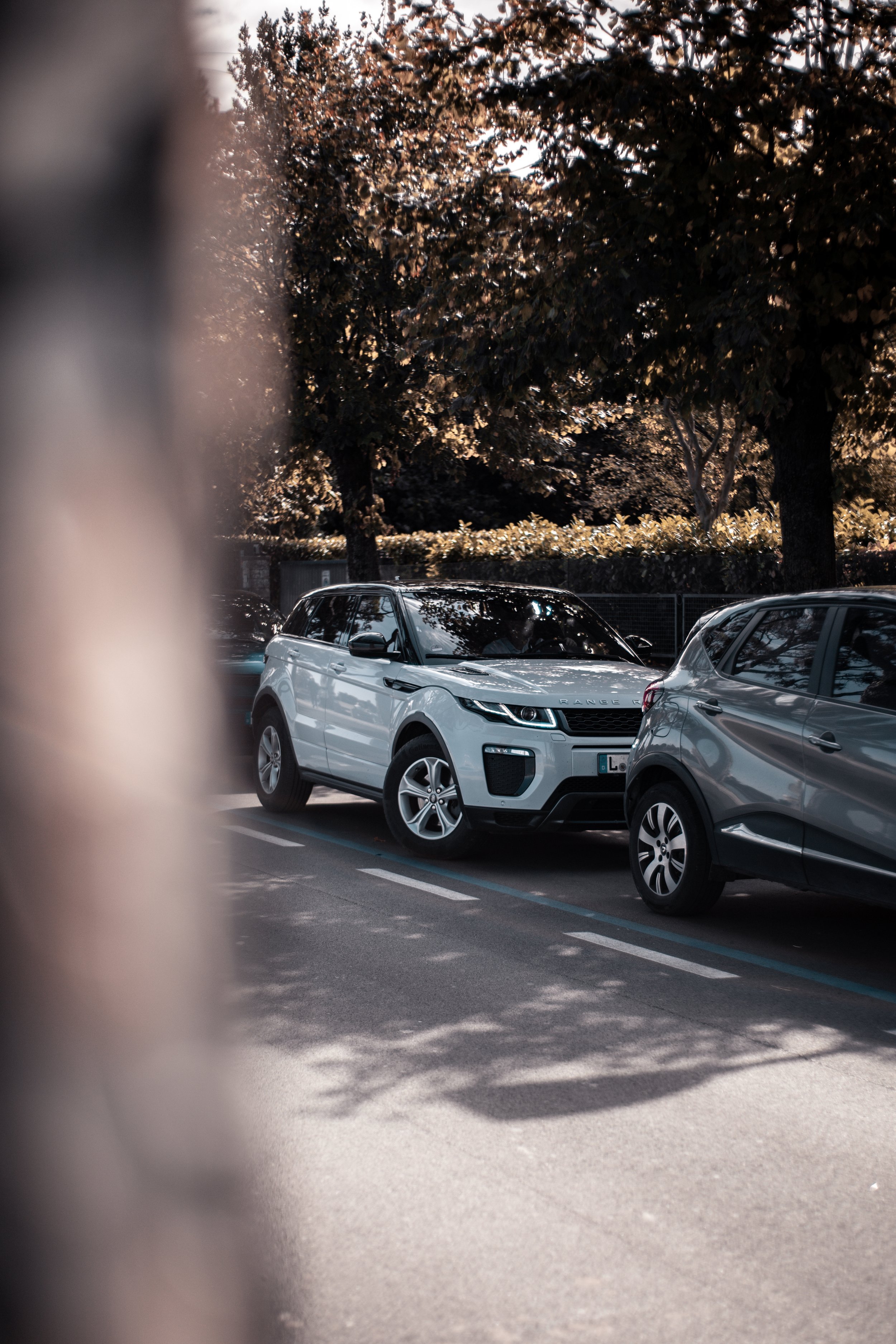 Ein Parkplatz mit mehreren geparkten Autos, im Hintergrund Bäume mit braunlaub, Blick durch eine Lücke zwischen einer weißen und einer grauen Autoseite im Vordergrund.