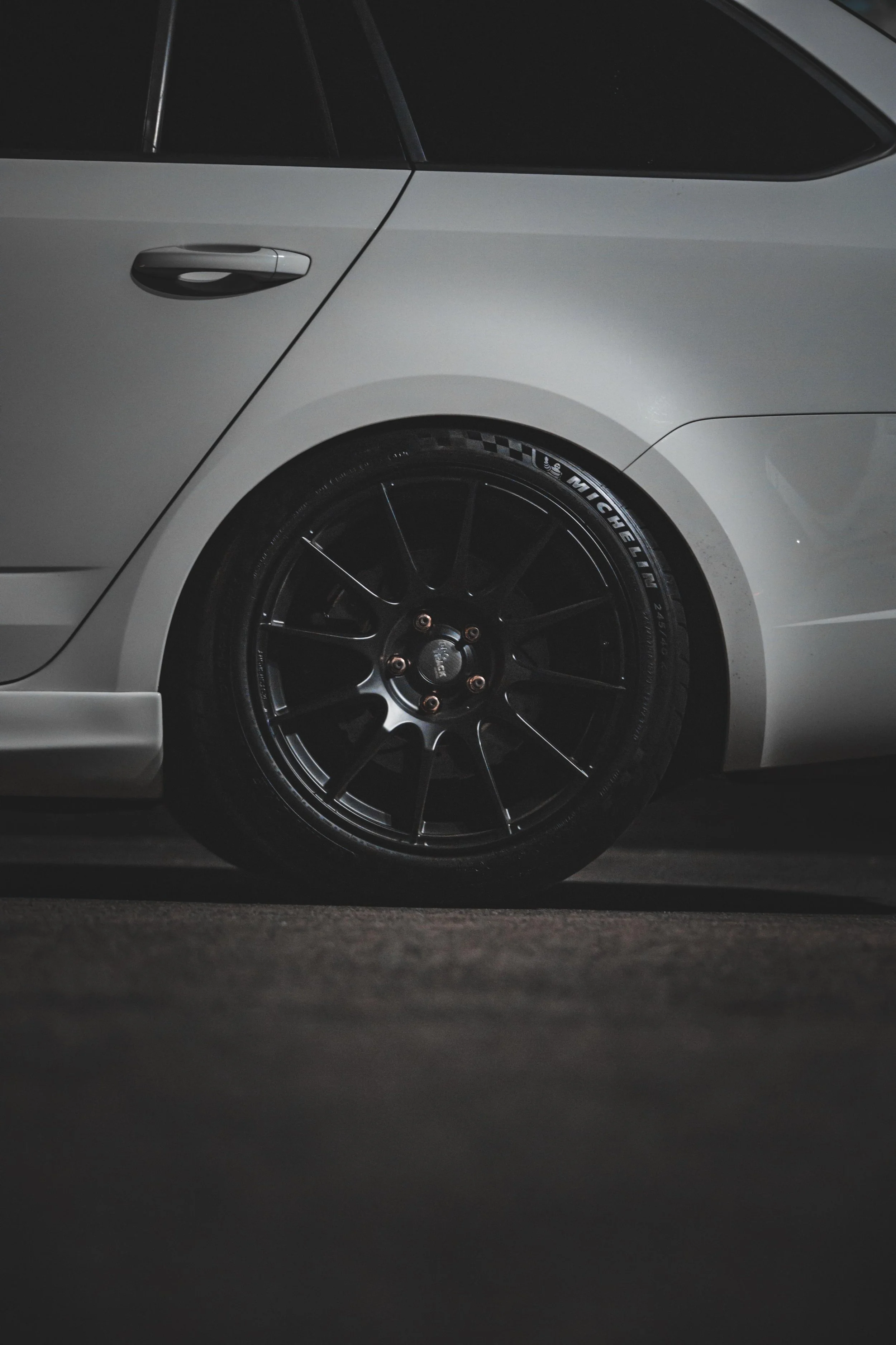 Nahaufnahme der hinteren Seite eines weißen Autos, Fokus auf das Rad mit schwarzen Felgen und Michelin-Reifen, im Dunkeln fotografiert.