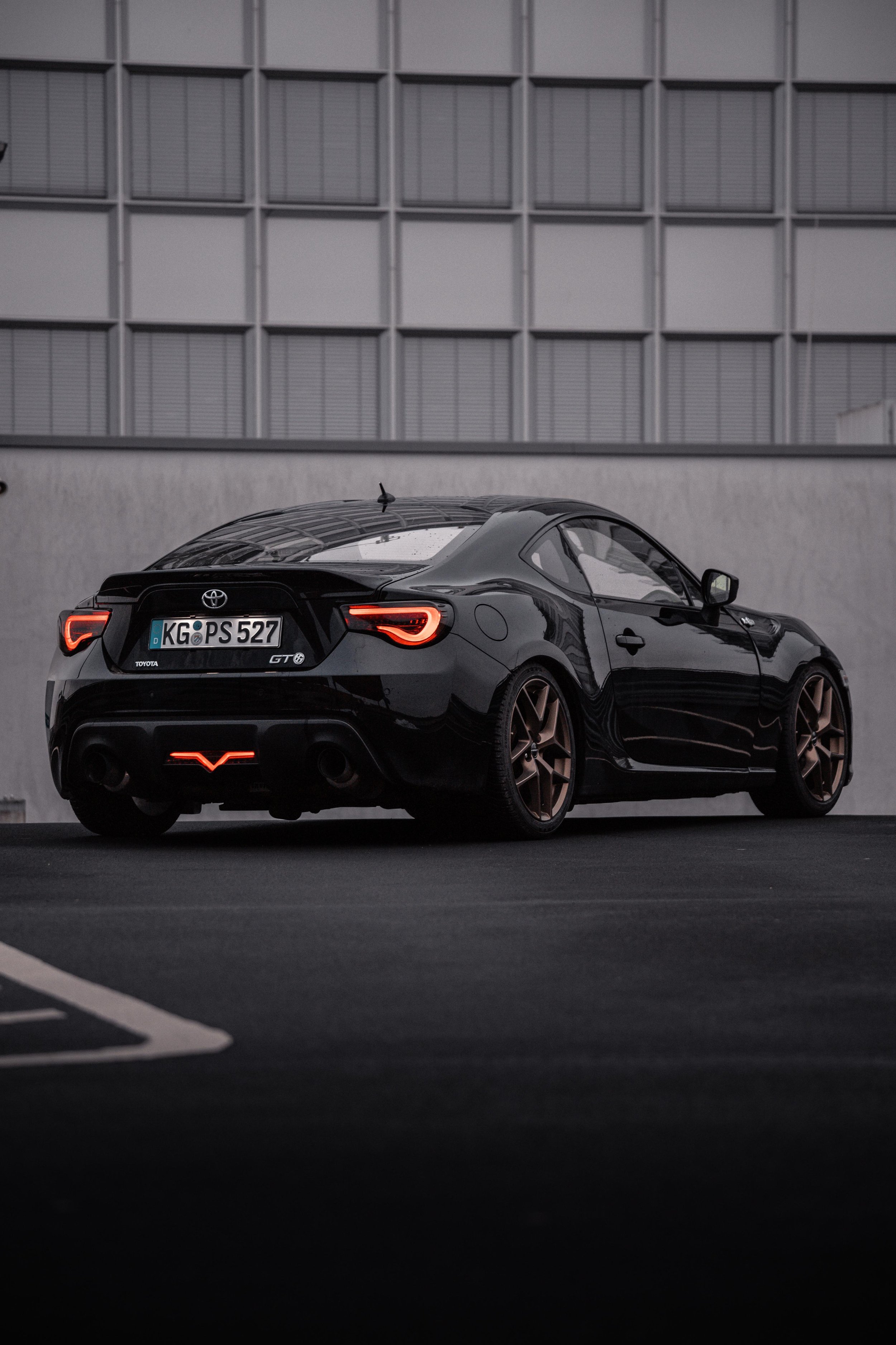 Schwarzer Toyota GT86 Auto parkt in einem dunklen Parkplatz vor einer grauen Wand mit einem modernen, mehrstöckigen Gebäude im Hintergrund. Es ist Dämmerung oder bewölkter Himmel, die Lichter des Autos sind eingeschaltet.