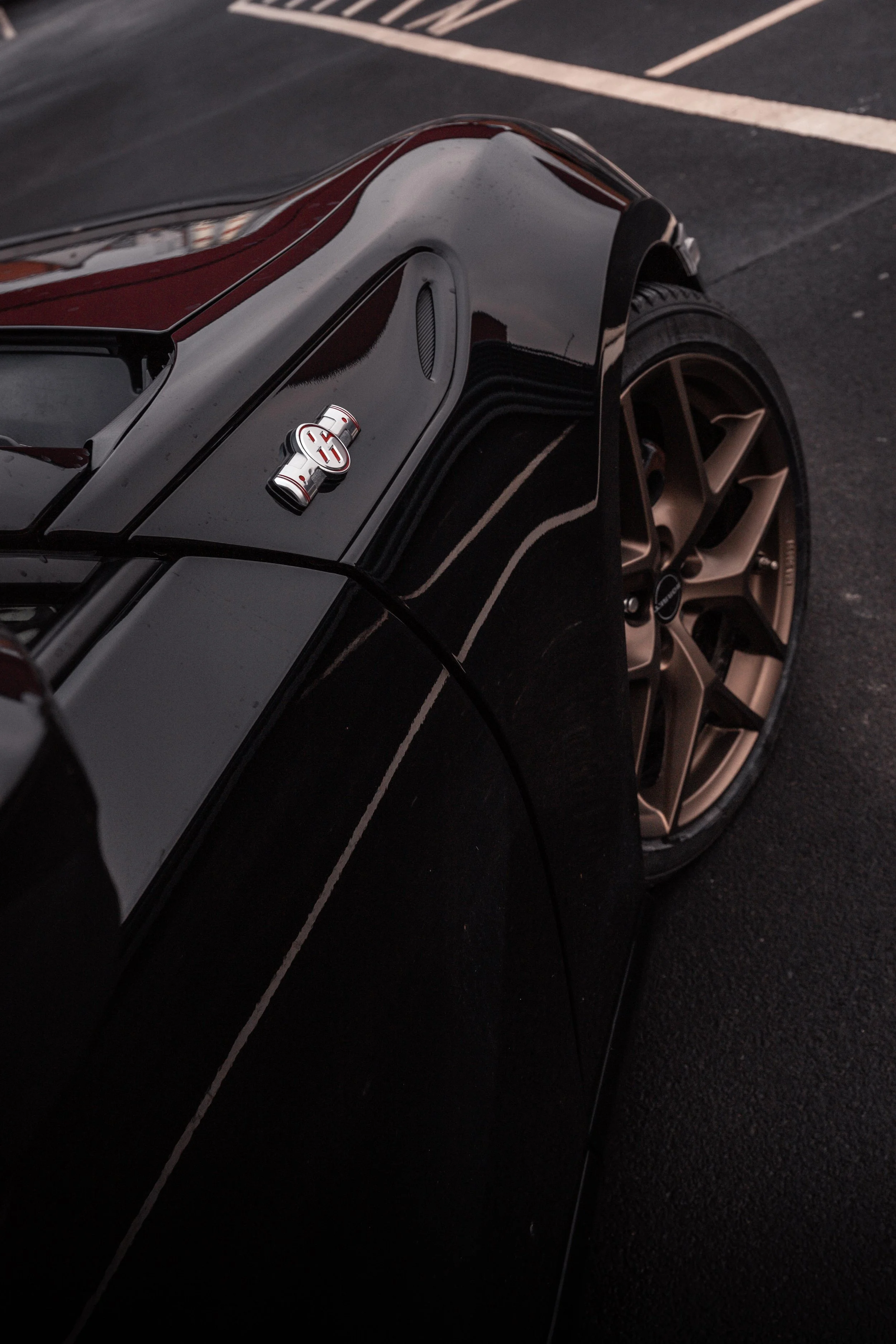 Nahaufnahme eines schwarzen Autos mit goldenen Felgen in einer Parklücke auf Asphalt, mit einem Chrome-Emblem auf der Motorhaube.