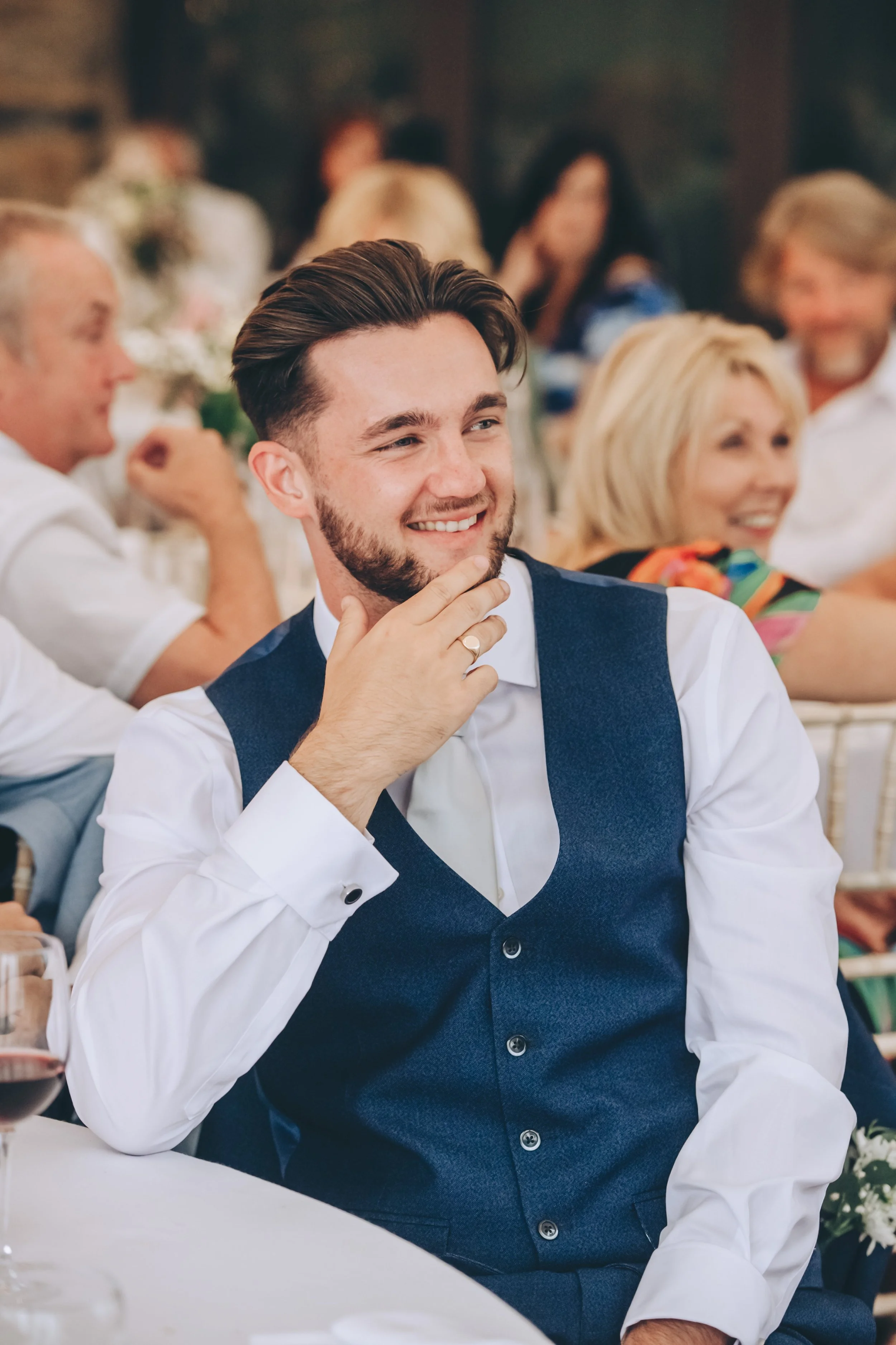 A young man with dark hair, beard, and light skin smiling and touching his chin with his hand, seated at a formal event with people in the background.