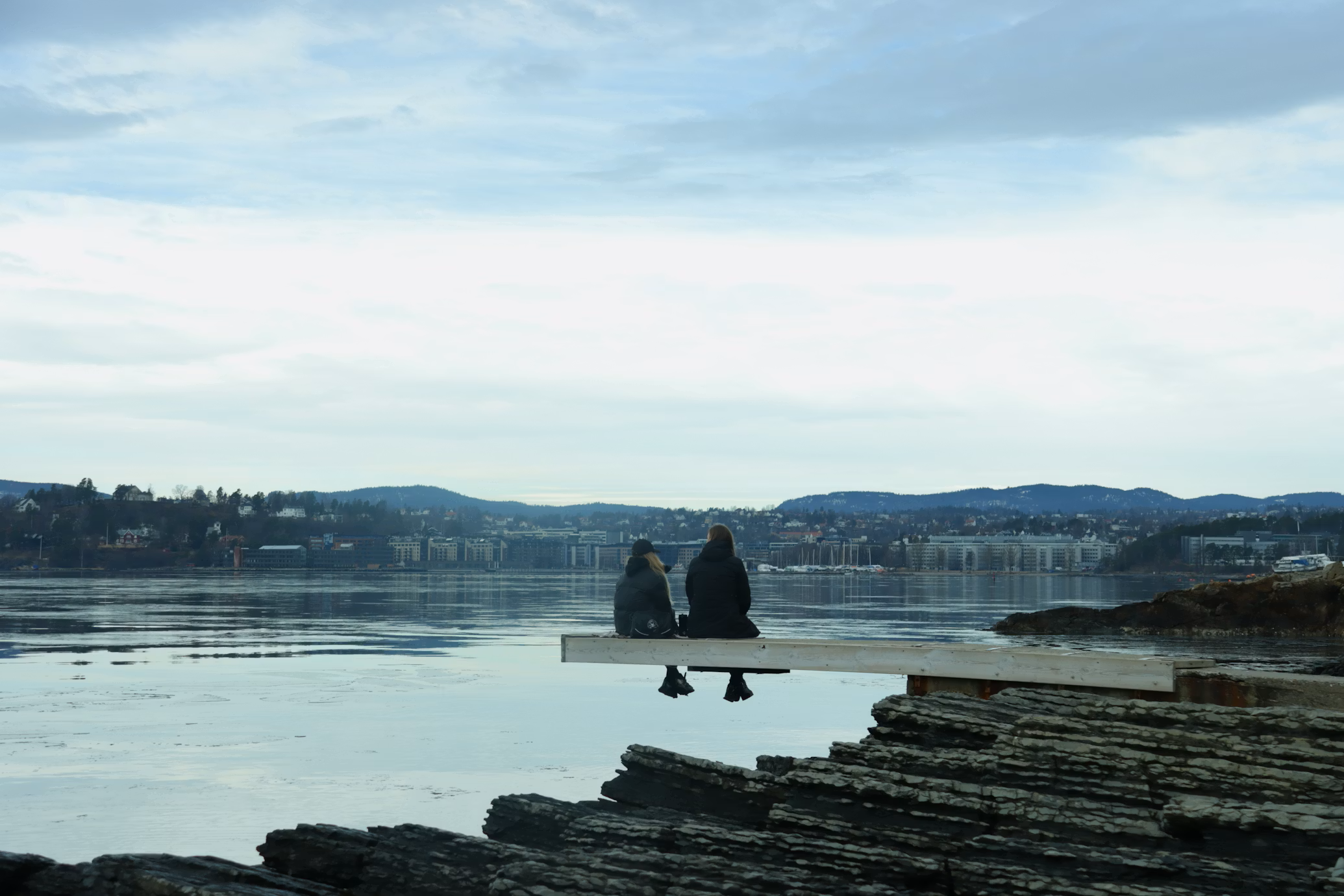 Two people sitting on a dock by the water, facing the city and hills in the distance on a cloudy day.