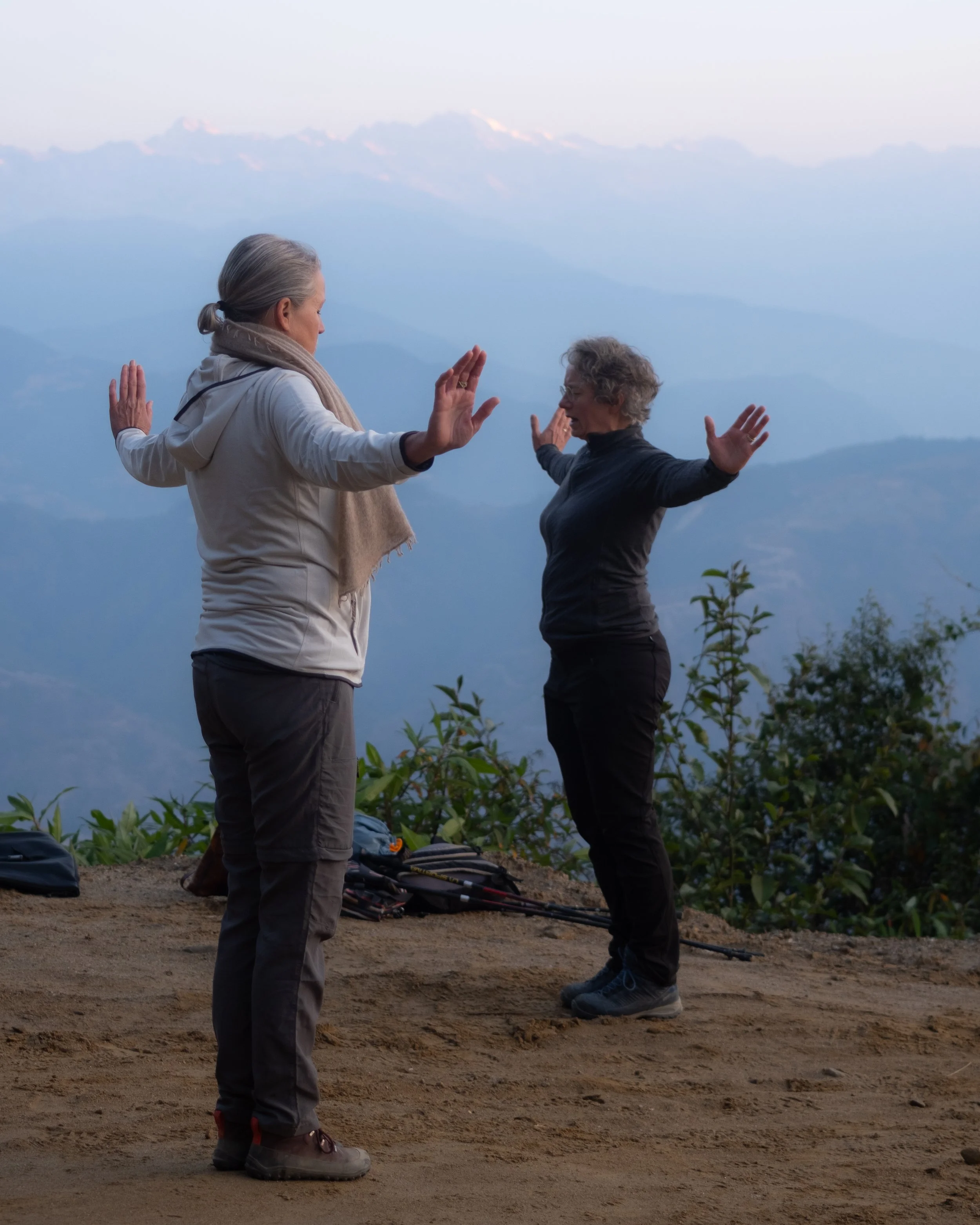 Twee vrouwen oefenen tai chi op een bergpad in de vroege ochtend, omringd door lage planten en met een berg en de hemel op de achtergrond.