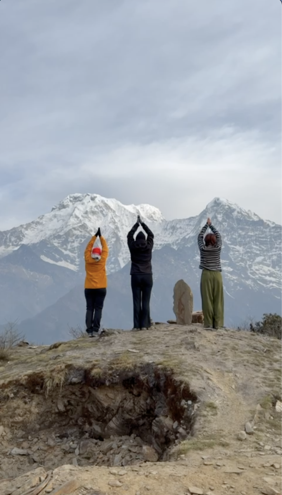 Drie mensen staan op een bergtop met sneeuwbedekte bergpieken op de achtergrond, alle drie maken ze een yogahouding met hun handen boven hun hoofd in een driedubbel oefening.