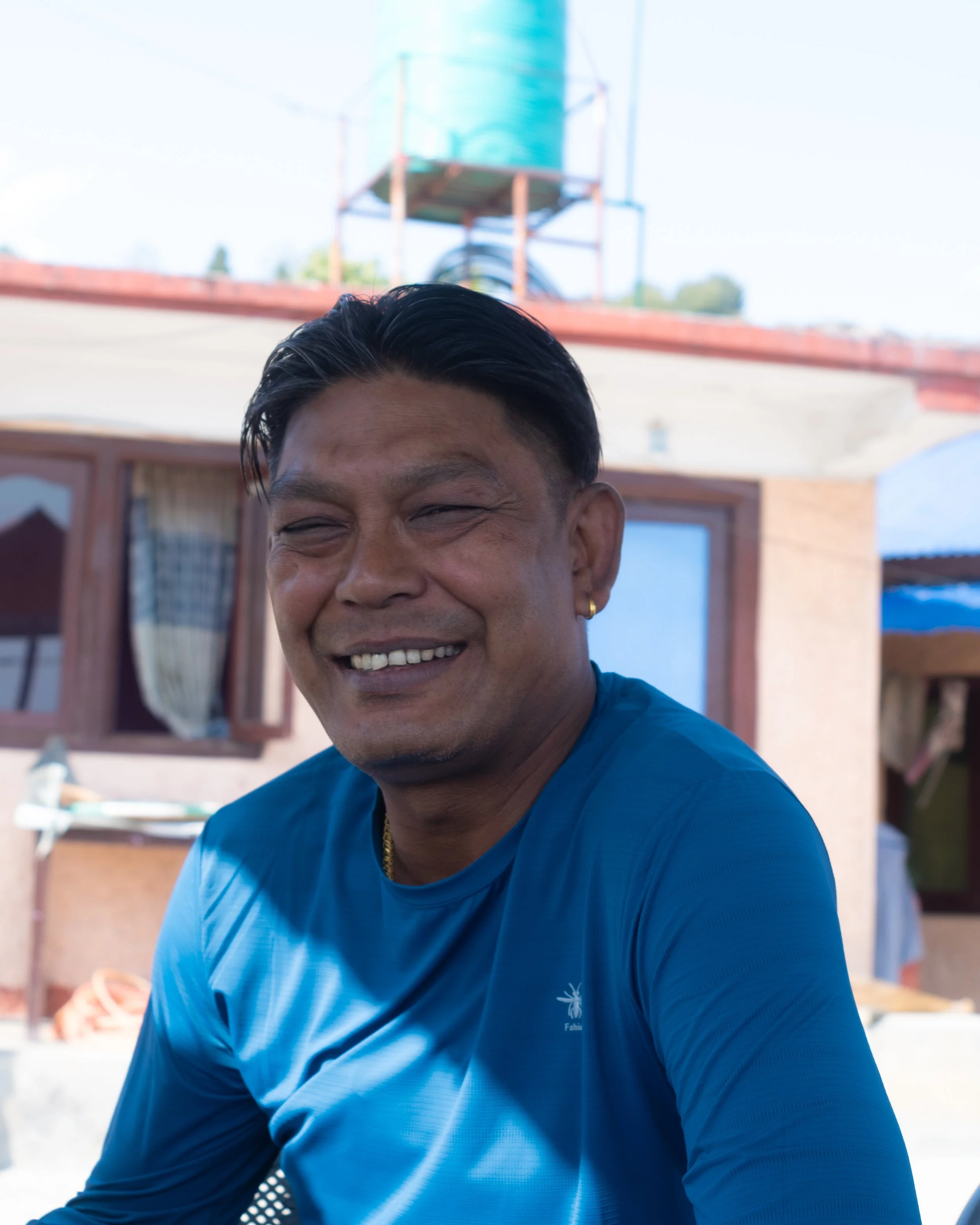 Lachende man met kort zwart haar, lichte huid, dragen een blauwe T-shirt met logo, buiten in een zonnige omgeving met een waterreservoir en huis op de achtergrond.
