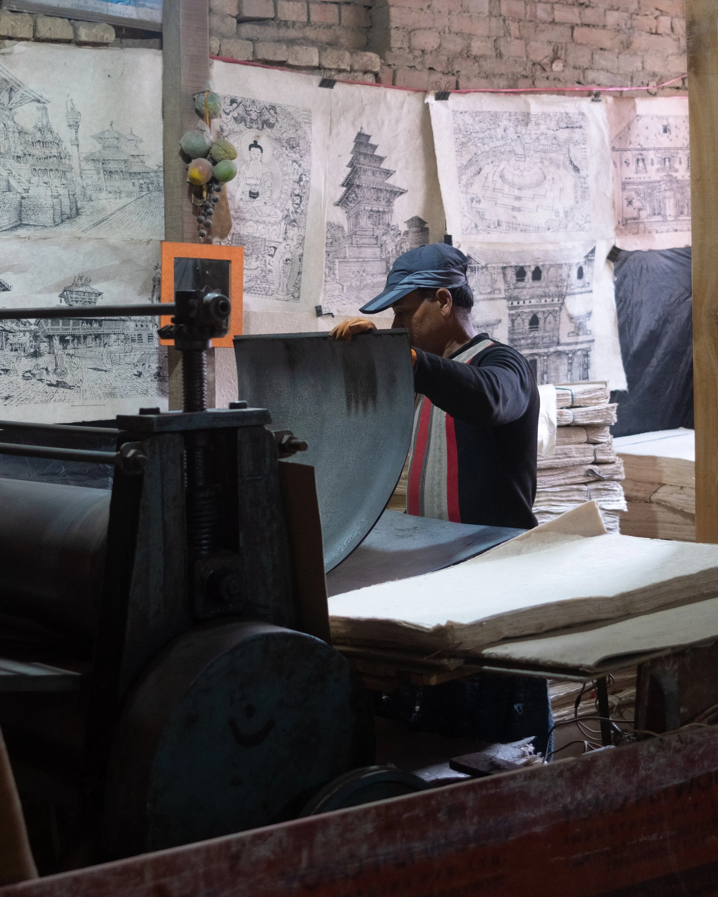 Een man met een pet en een schort werkt in een houtwerkplaats, omringd door houtblokken en tekeningen van gebouwen en monumenten die aan de muur hangen.
