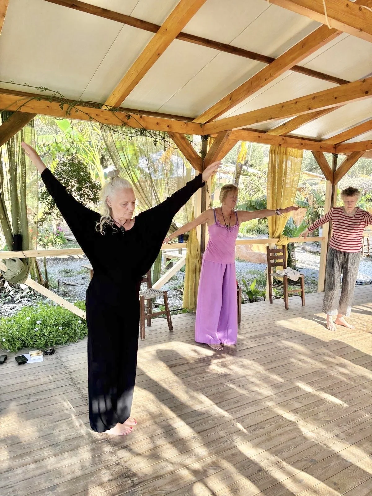Drie vrouwen die yoga of meditatie oefeningen doen onder een houten dak in een tuin met groene planten.