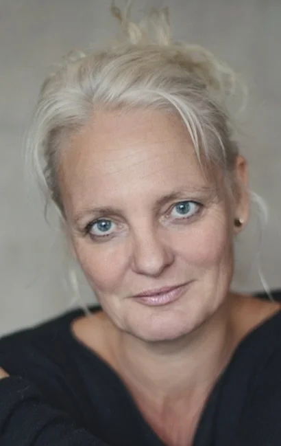 Een close-up portret van een vrouw met blonde, licht golvende haar en blauwe ogen, gekleed in een zwart top.