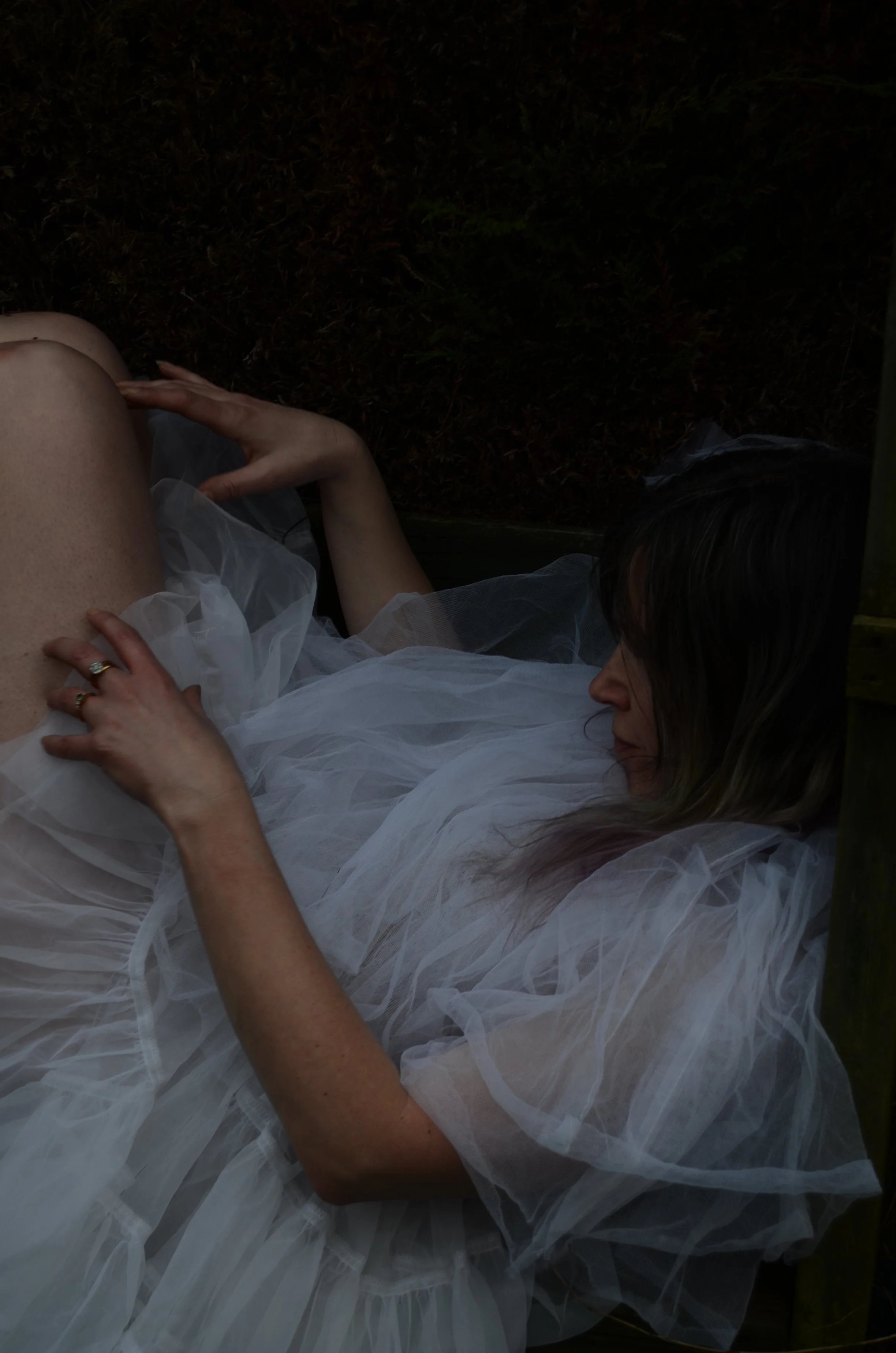 A woman in a white dress with sheer fabric lying down on her side on a dark surface, with her hand resting on her thigh and her head turned to the side.
