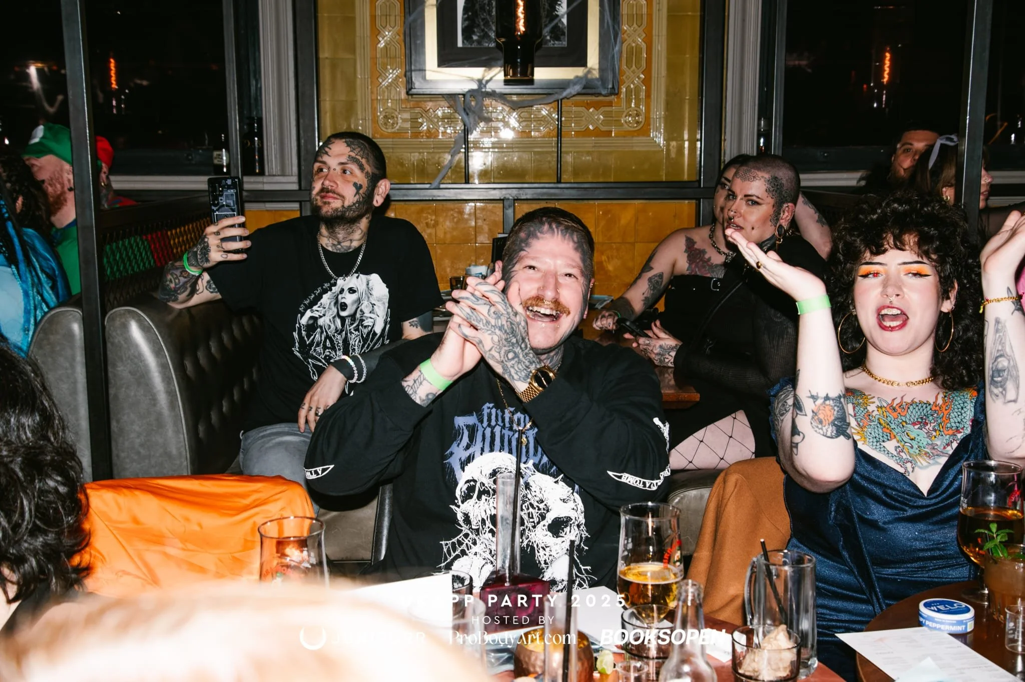 Group of diverse people celebrating at a party, with drinks on the table and vibrant tattoos, some holding glasses or making gestures, in a lively indoor setting.