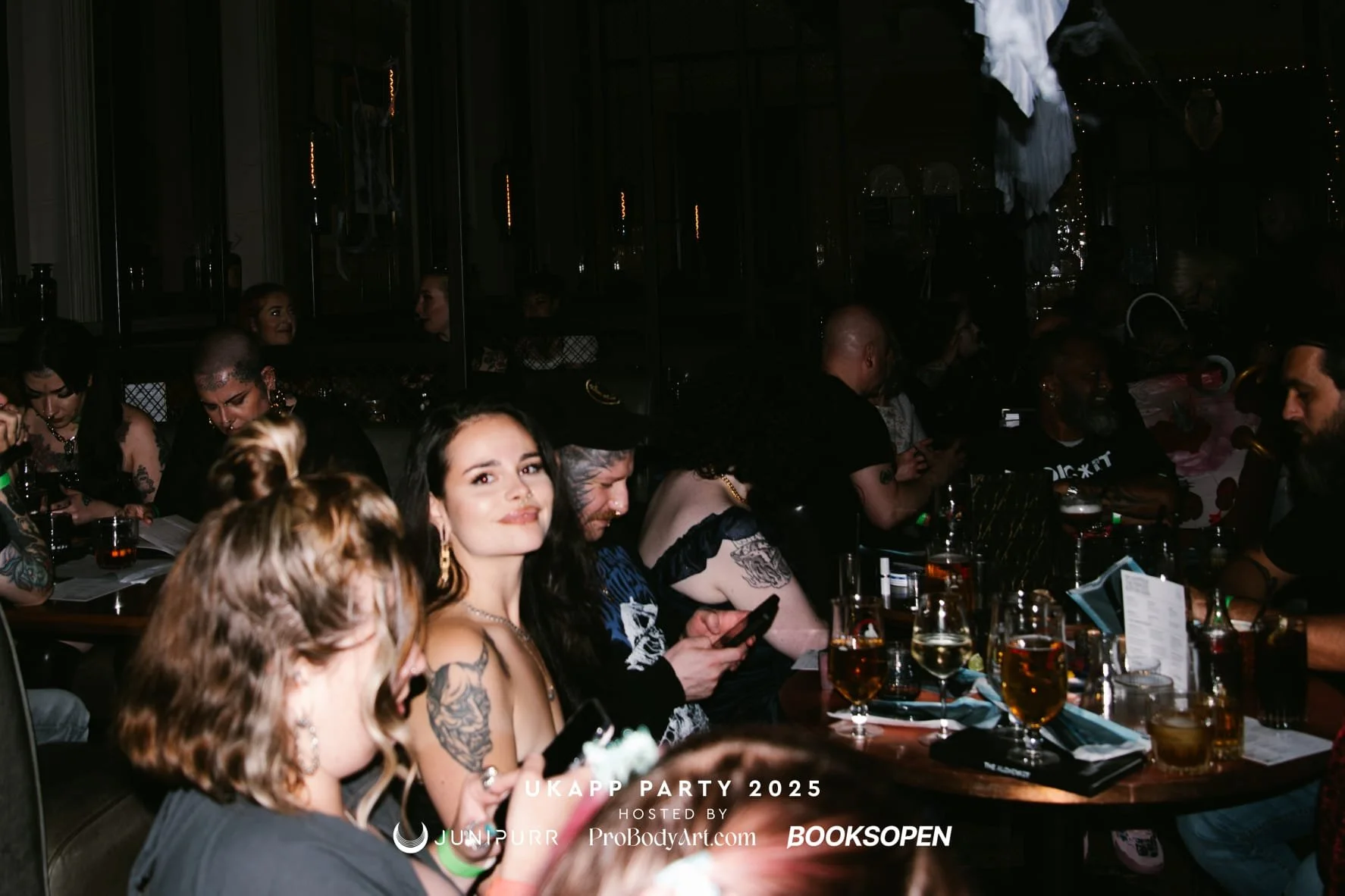 People sitting at a long dinner table during a party, smiling and looking at the camera, with drinks and menus in front of them, with a dark background and some decorative lights.