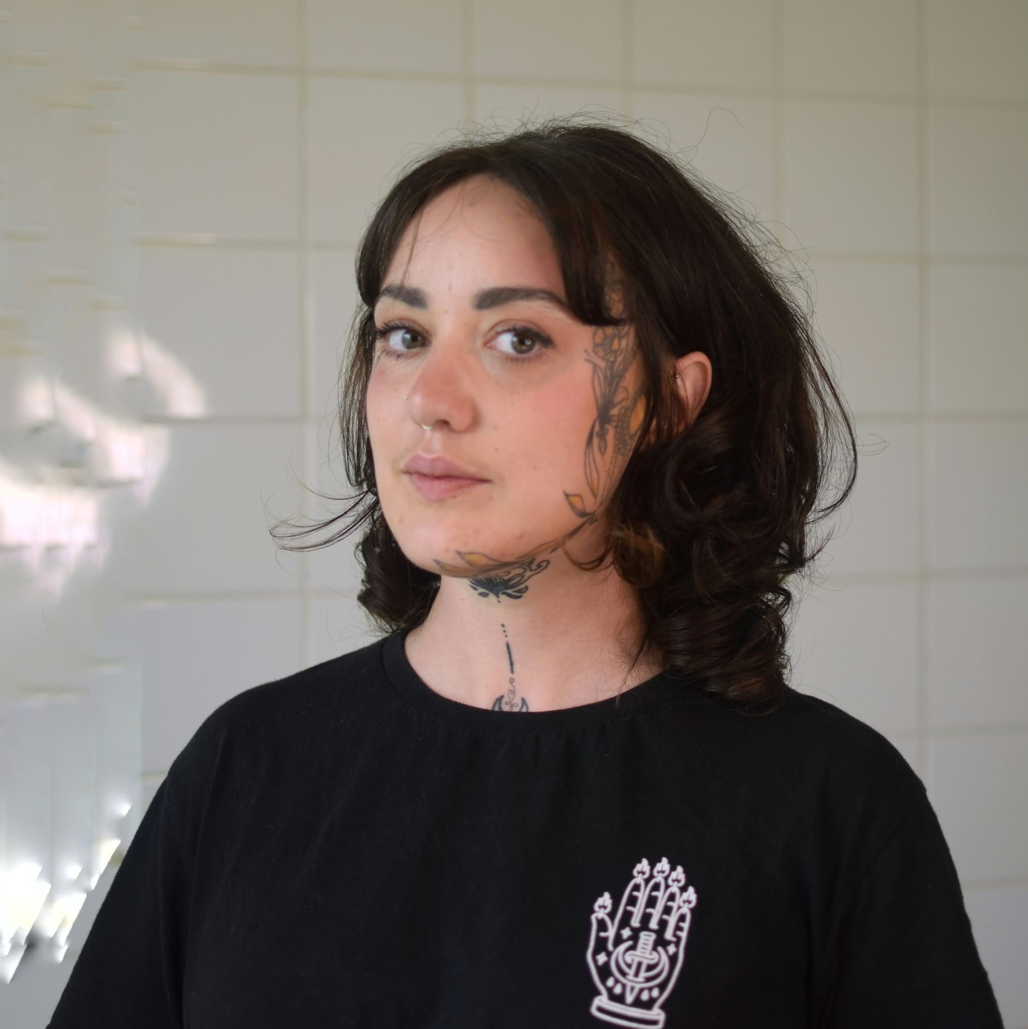 A woman with dark hair and tattoos on her face and neck, wearing a black shirt with a white graphic design, stands in front of a light-colored tiled wall.