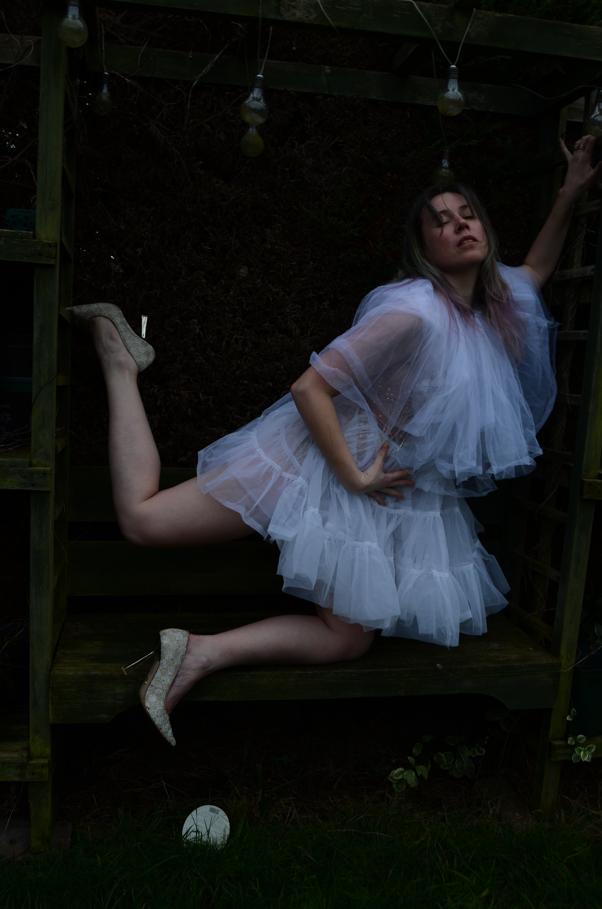 A woman wearing a white tulle dress and glittery high heels, posing in a precarious position inside a wooden frame outdoor setting with string lights overhead and grass at the bottom.