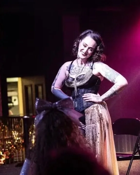 A woman on stage posing with a smile, dressed in vintage-style clothing and tattoos visible on her arms, with a person in front watching.