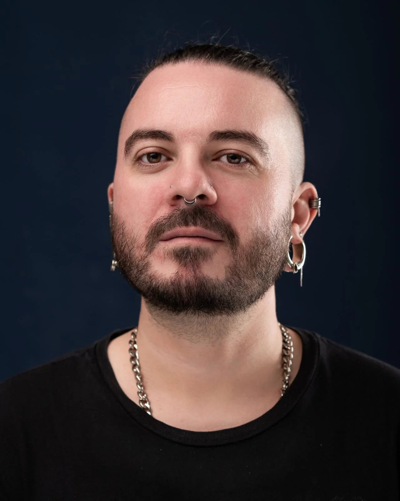 Close-up portrait of a man with multiple facial piercings, earrings, and a chain necklace, wearing a black shirt, against a dark background.