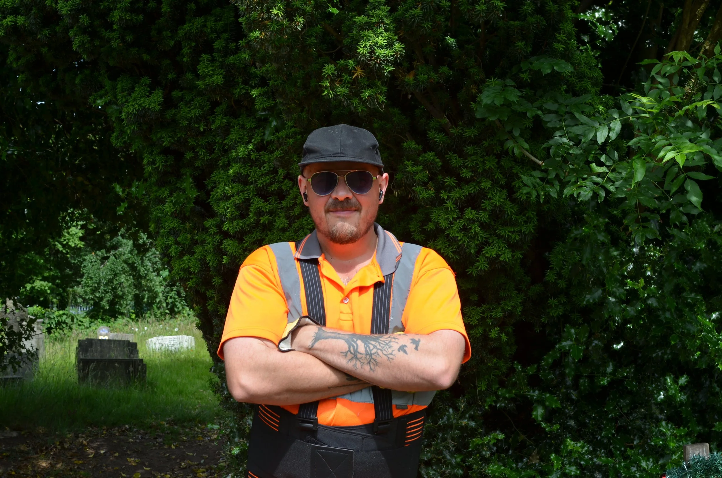 A man with sunglasses and a black cap, wearing an orange safety shirt, standing outdoors with arms crossed in front of a large green leafy bush.