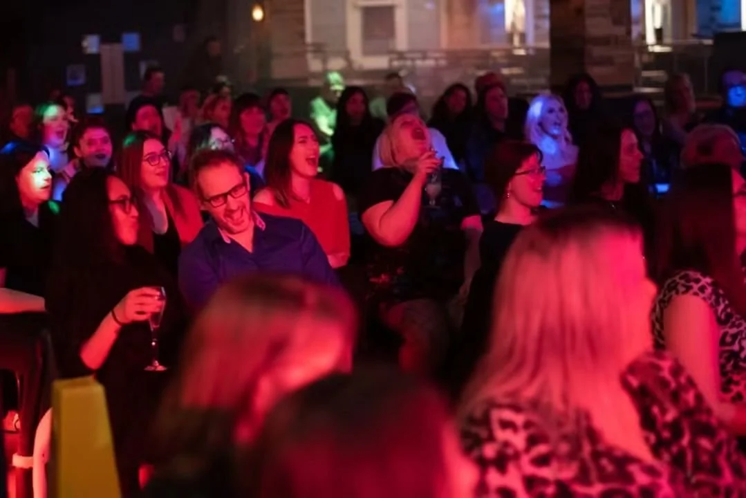 A group of women at a cabaret burlesque show live event, sitting in an audience reacting, laughing, and enjoying the performance with colorful stage lighting around them.