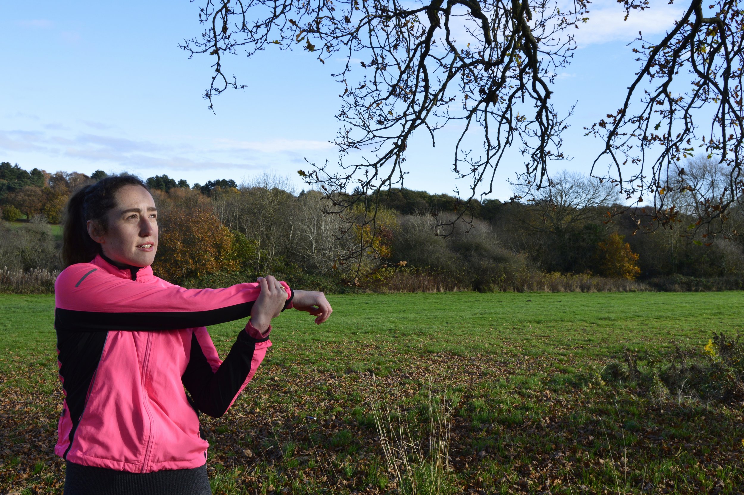 A woman in a pink jacket stretching in a park with trees and a hill in the background.