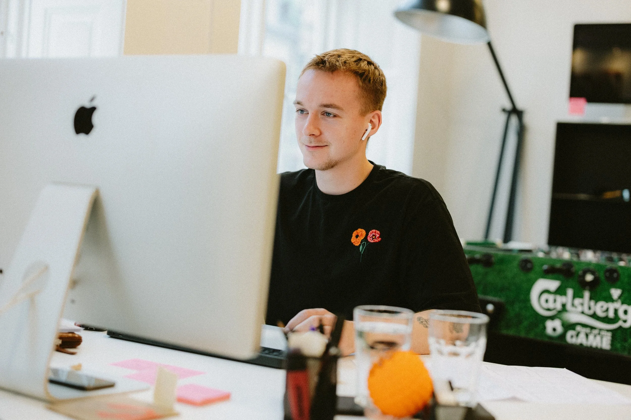 A young man working at a desk with Apple computer, wearing wireless earbuds and a black t-shirt with a floral embroidery, in a bright room with windows and a green Carlsberg Game sticker on a box in the background.
