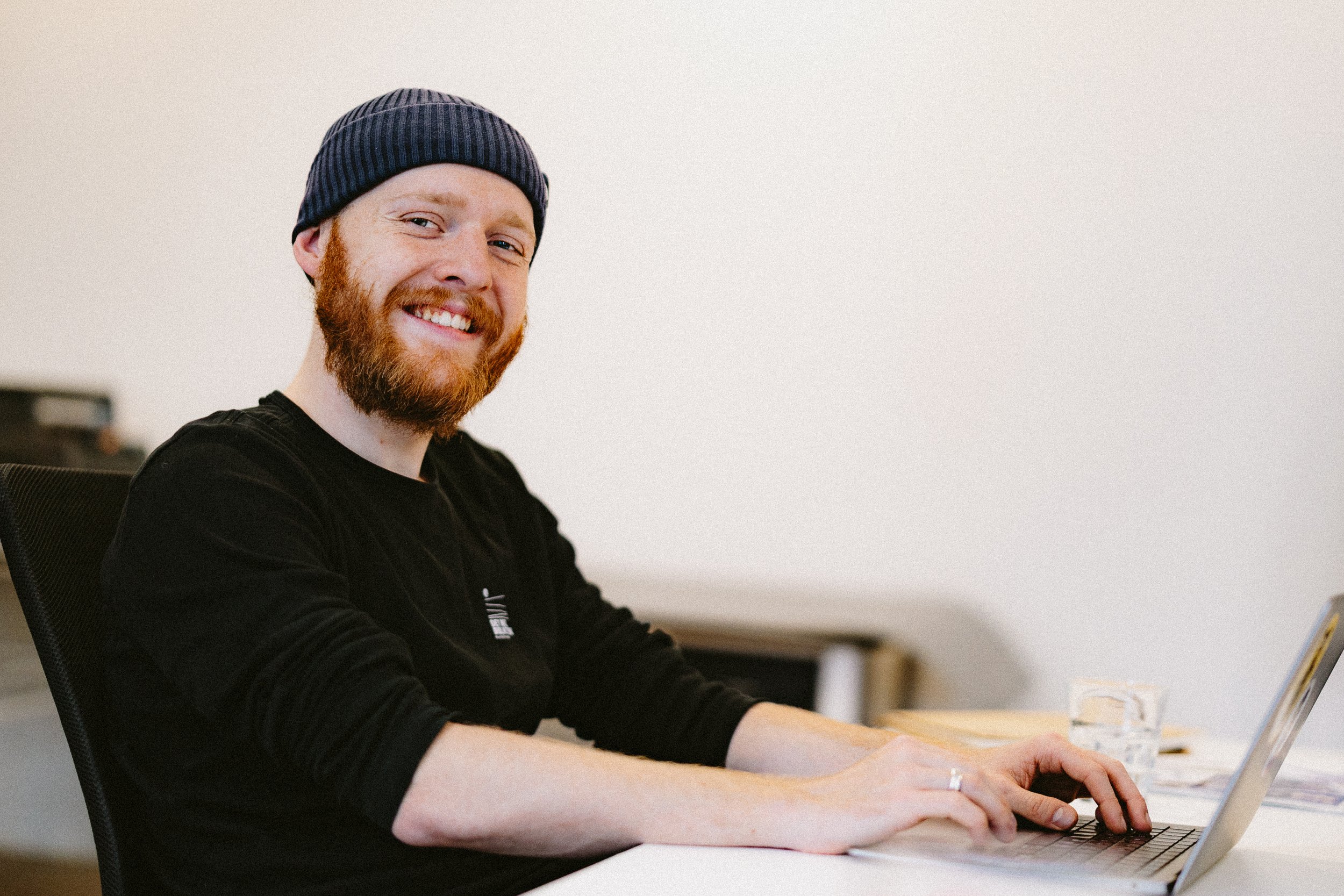 A smiling man with red hair, beard, and a beanie, sitting at a desk using a laptop.