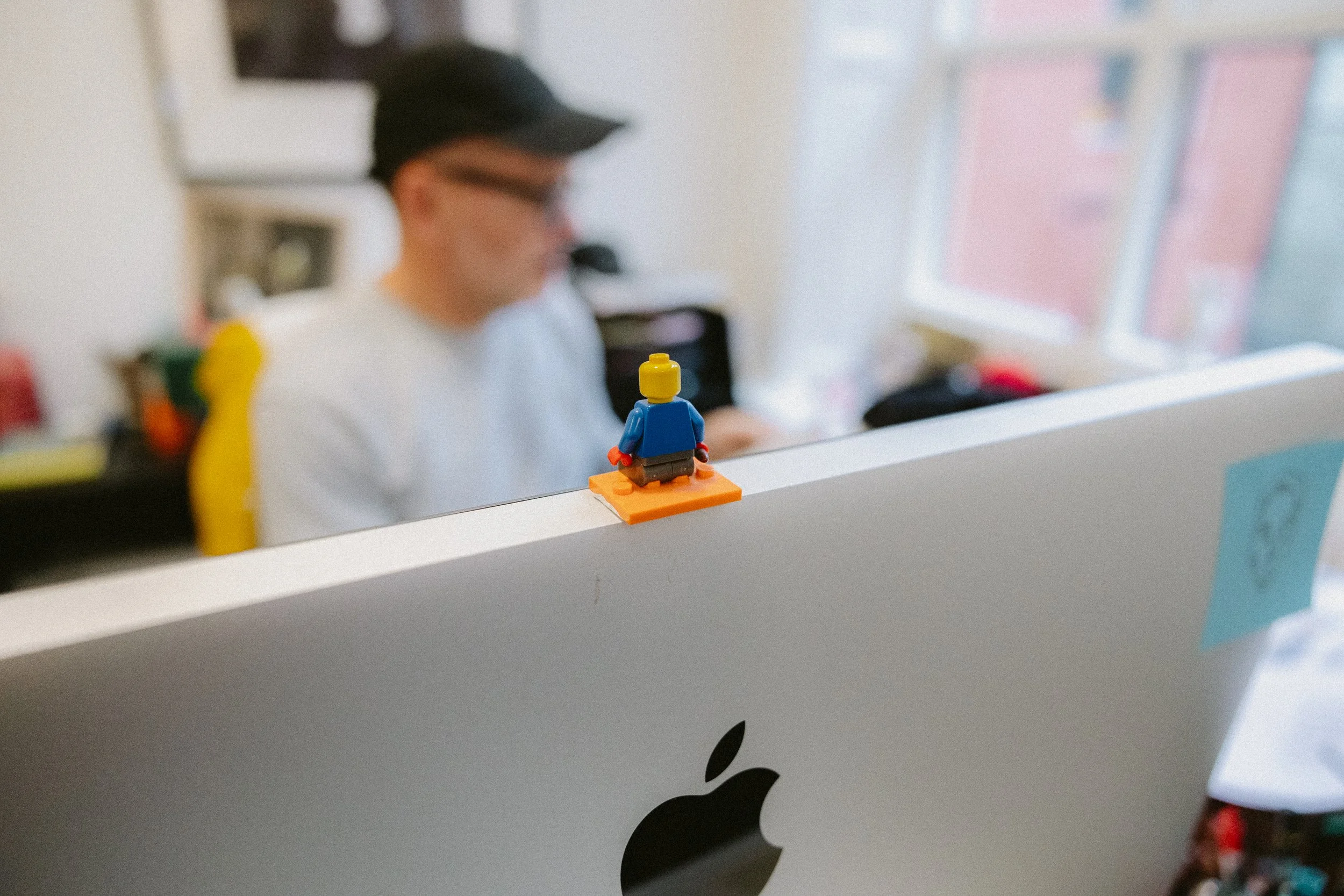 A small LEGO figure of a person with a yellow head, blue shirt, and black pants sits on an orange platform on top of an iMac computer, with a blurred person wearing a baseball cap and glasses in the background.