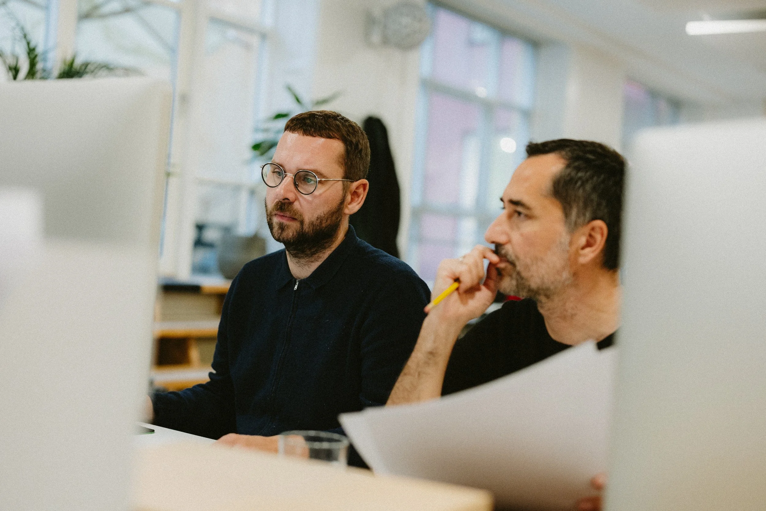 Two men working at a desk in an office, surrounded by computer monitors and office supplies, with windows and plants in the background.