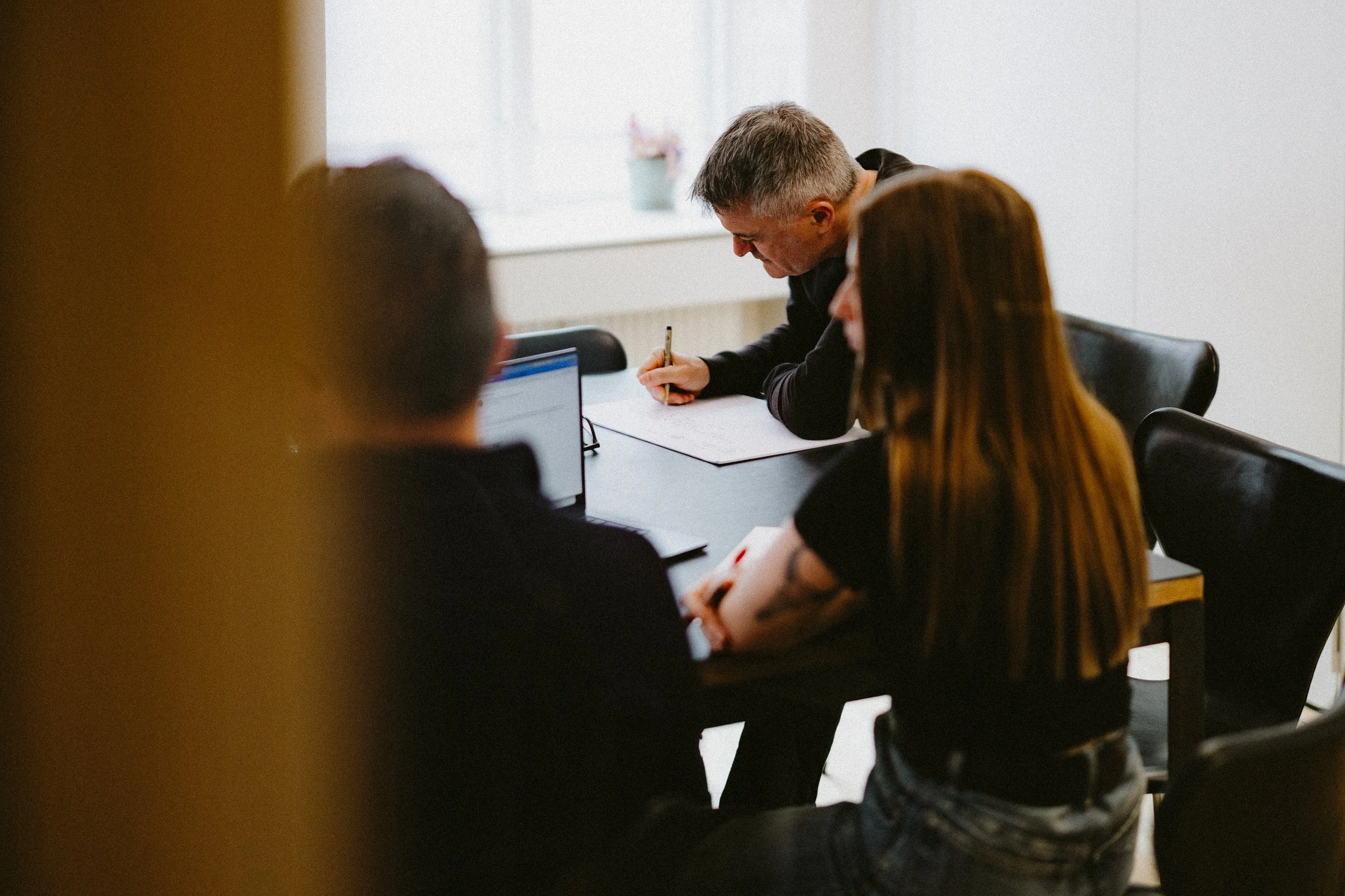 Three people sitting at a black table with one writing in a notebook, in a bright office with white walls and a window.