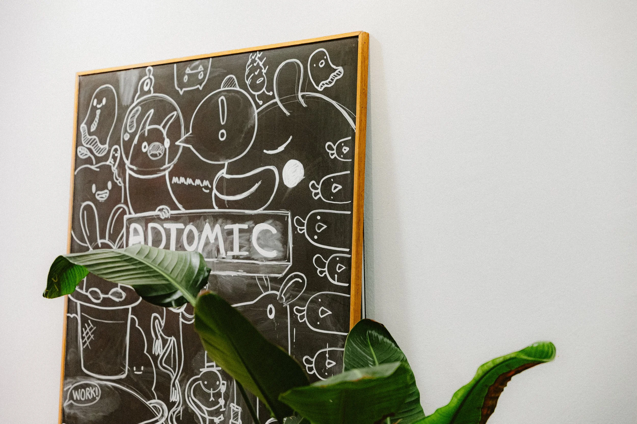 Black chalkboard with cartoon doodles and the word 'ADTOMIC' written on it, partially obscured by large green leaves of a plant in front.