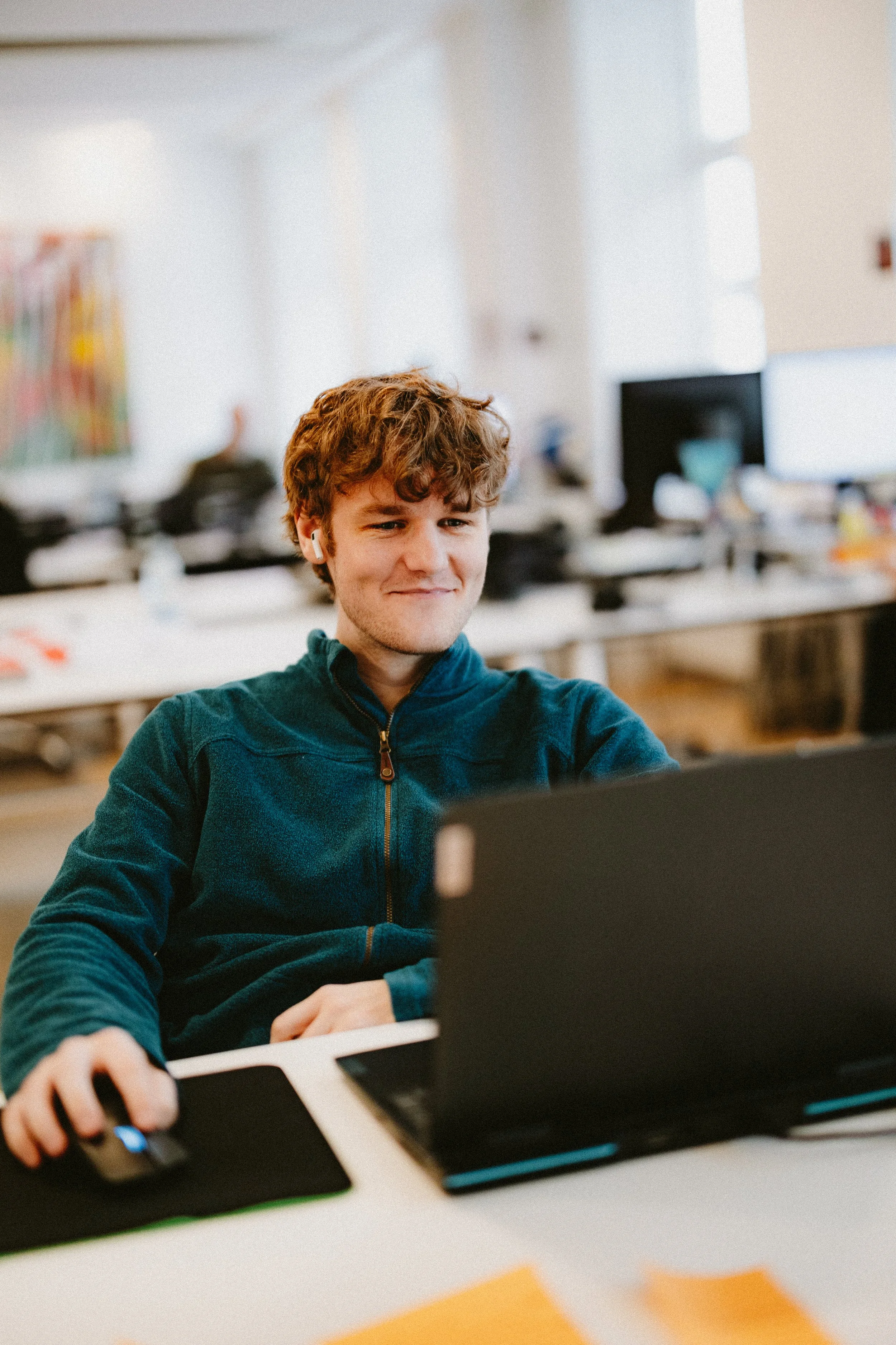 A young man with brown curly hair, wearing a dark teal zip-up hoodie, is sitting at a desk using a laptop and a computer mouse in an office setting.