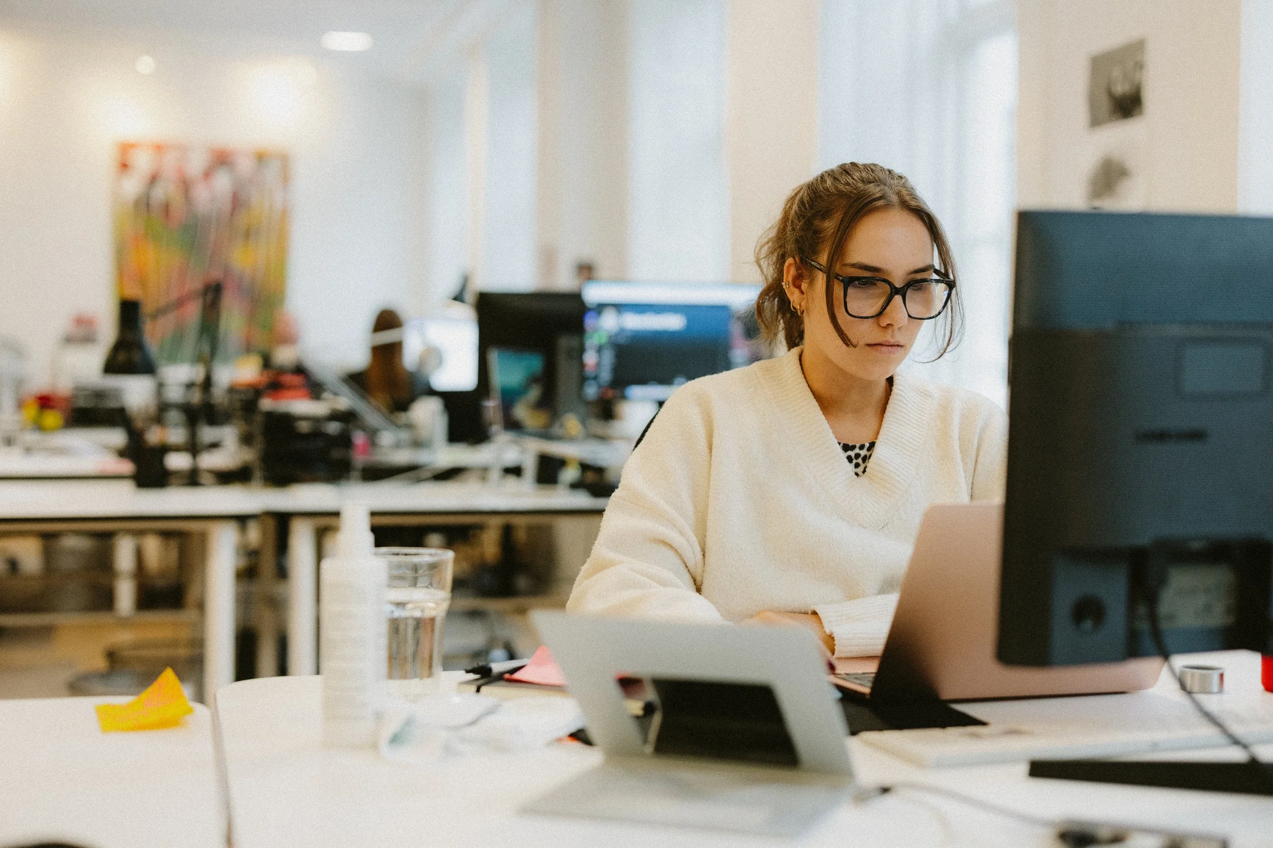Woman with glasses working at her desk in an office environment, surrounded by multiple computer monitors and office supplies.