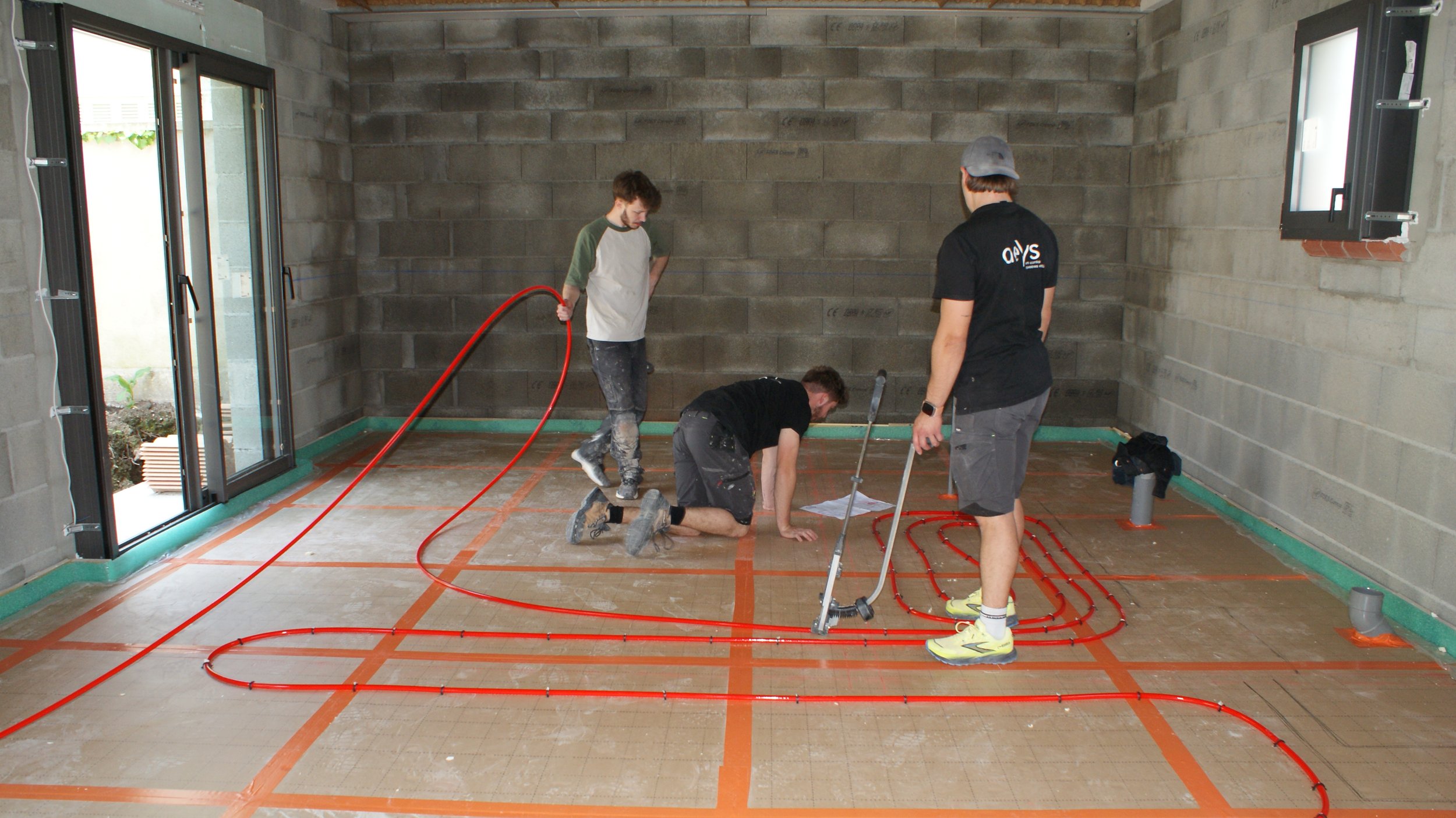 Quatre hommes travaillent dans une pièce en construction, installant un système de chauffage par le sol avec des tuyaux enroulés rouges sur le sol en béton. La pièce a des murs en parpaings, deux fenêtres ouvertes, et il y a des outils et des documents sur le sol.