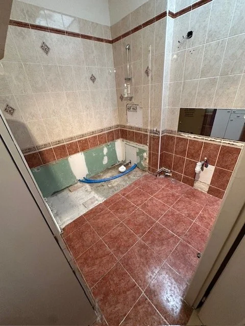Salle de bain en rénovation, avec carreaux de sol marron et murs partiellement carrelés, absence d'équipements sanitaires installés, plomberie visible.