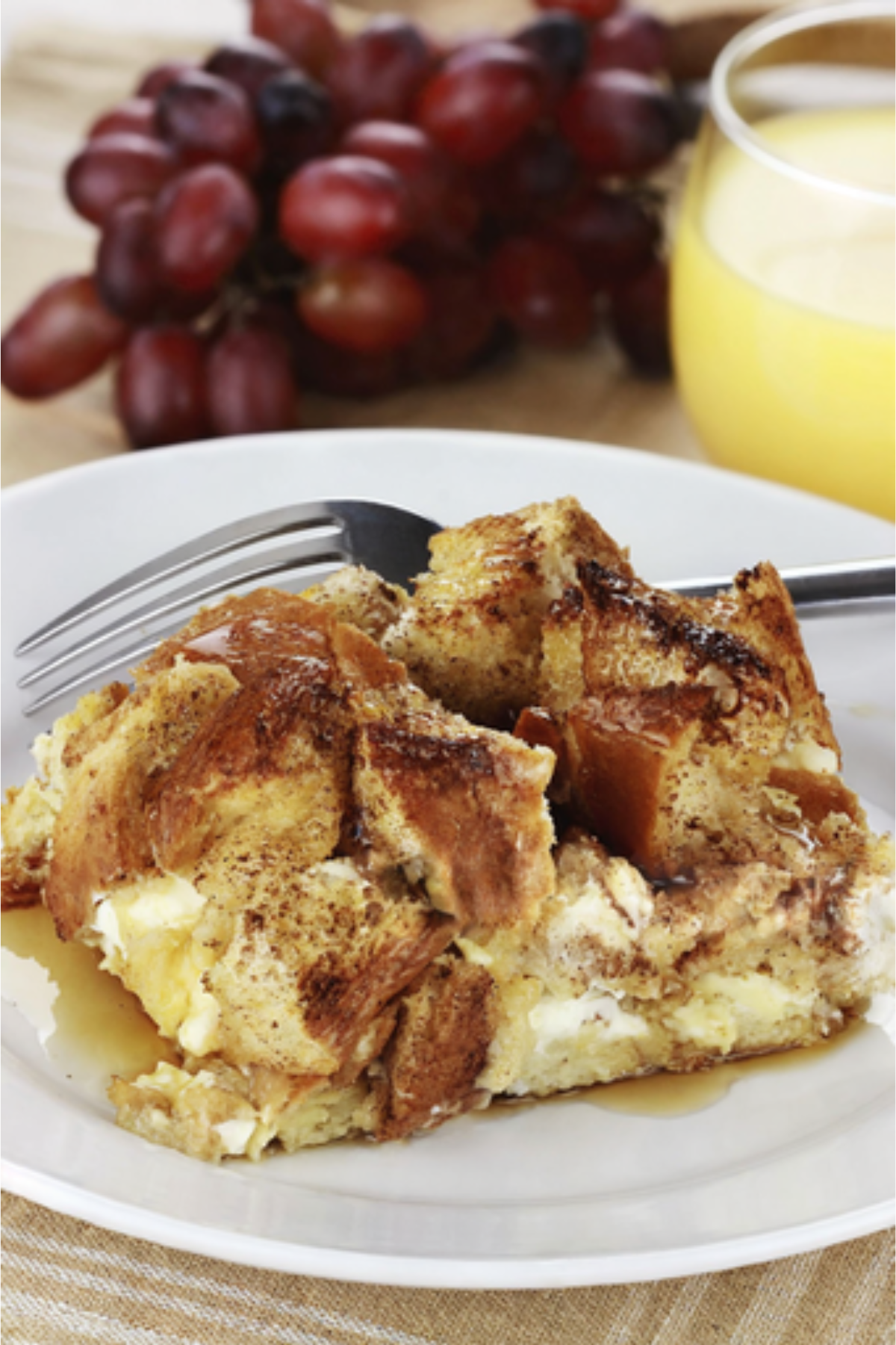 A slice of French toast casserole on a white plate, with a fork, with grapes and a glass of orange juice in the background.