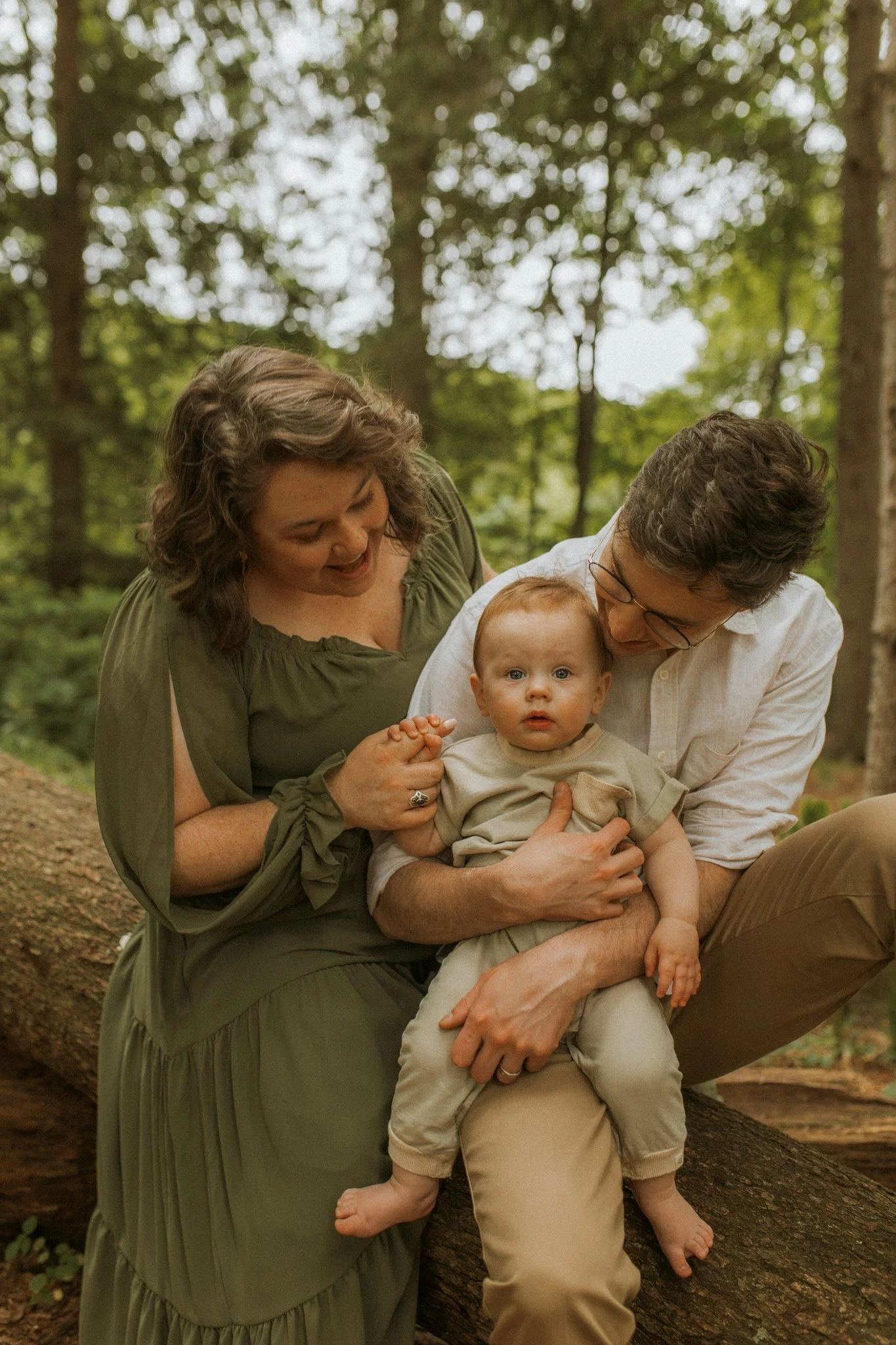A family of three, including a woman, man, and a young child, sitting together on a large fallen tree trunk in a wooded forest area.