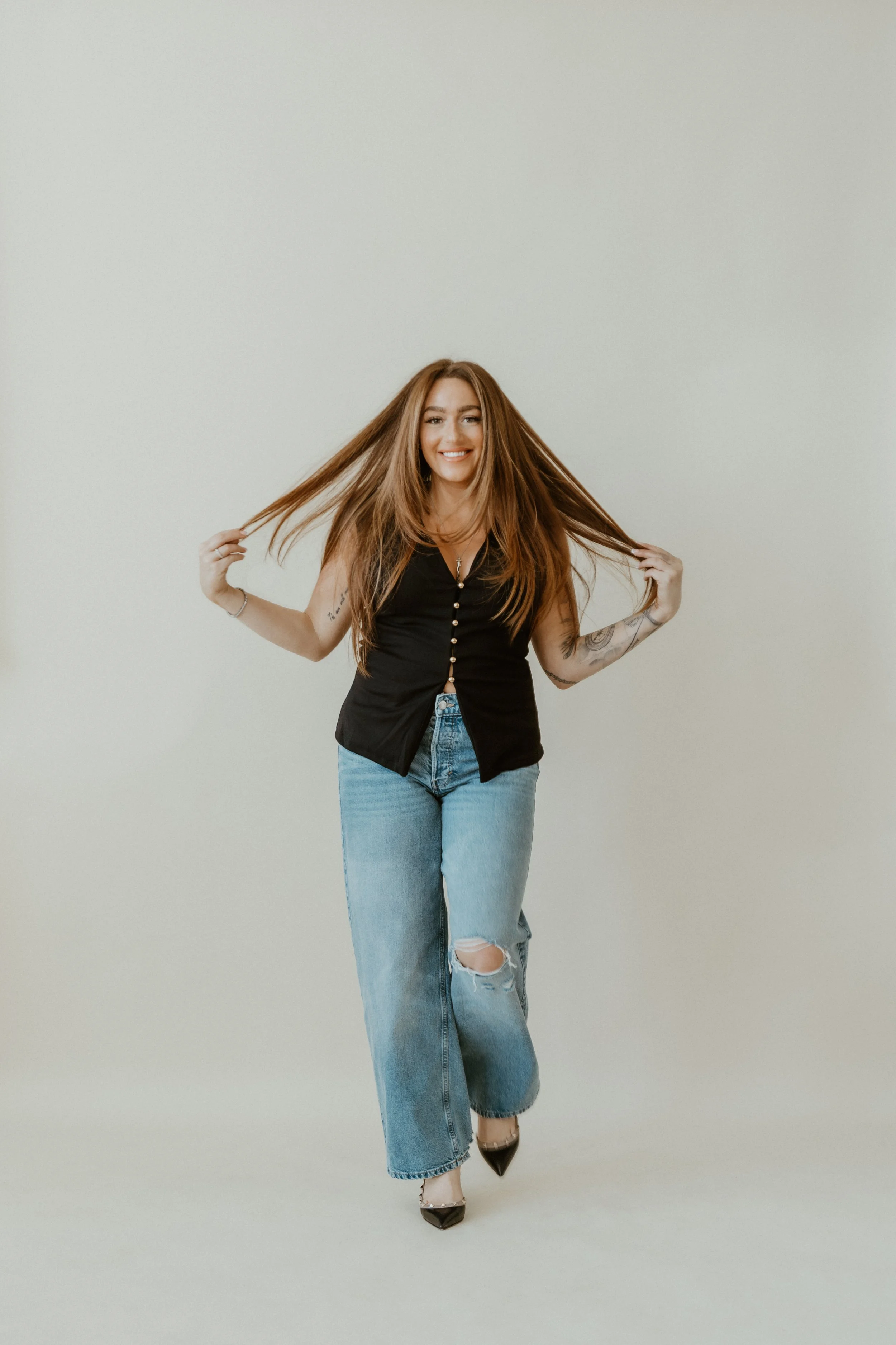 A woman with long, reddish-brown hair and tattoos on her arms, smiling, standing against a plain, light-colored background, wearing a black sleeveless top, light blue ripped jeans, and black pointed shoes. She is holding her hair with both hands.