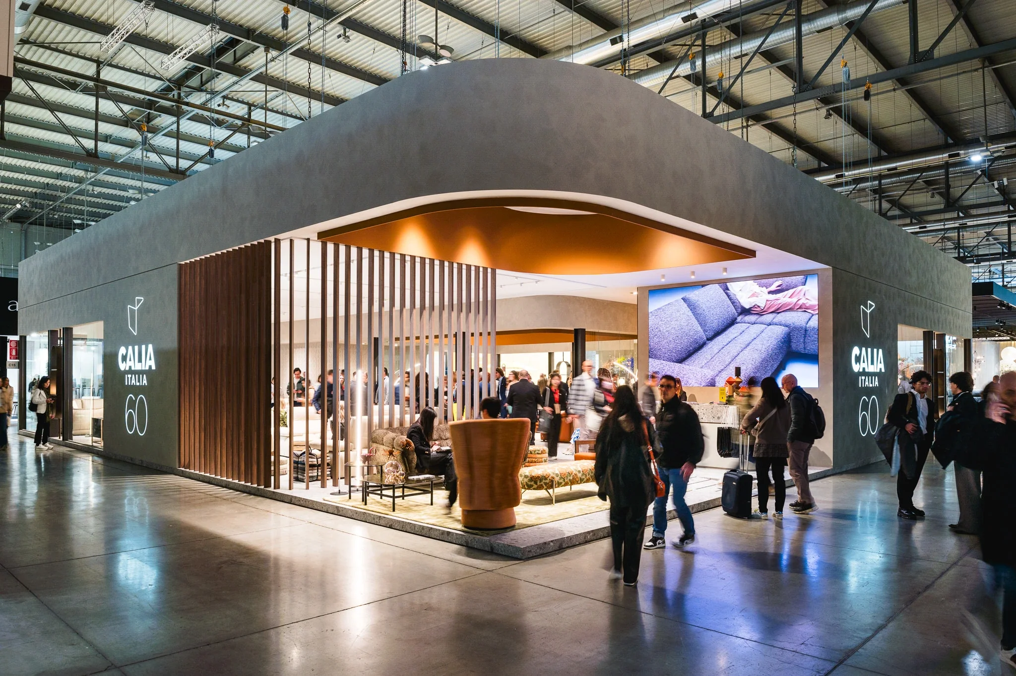 Stand fieristico di Calia Italia con persone che visitano, arredamento di divani e poltrone, grande schermo digitale, struttura moderna e luminosa in un ambiente industriale