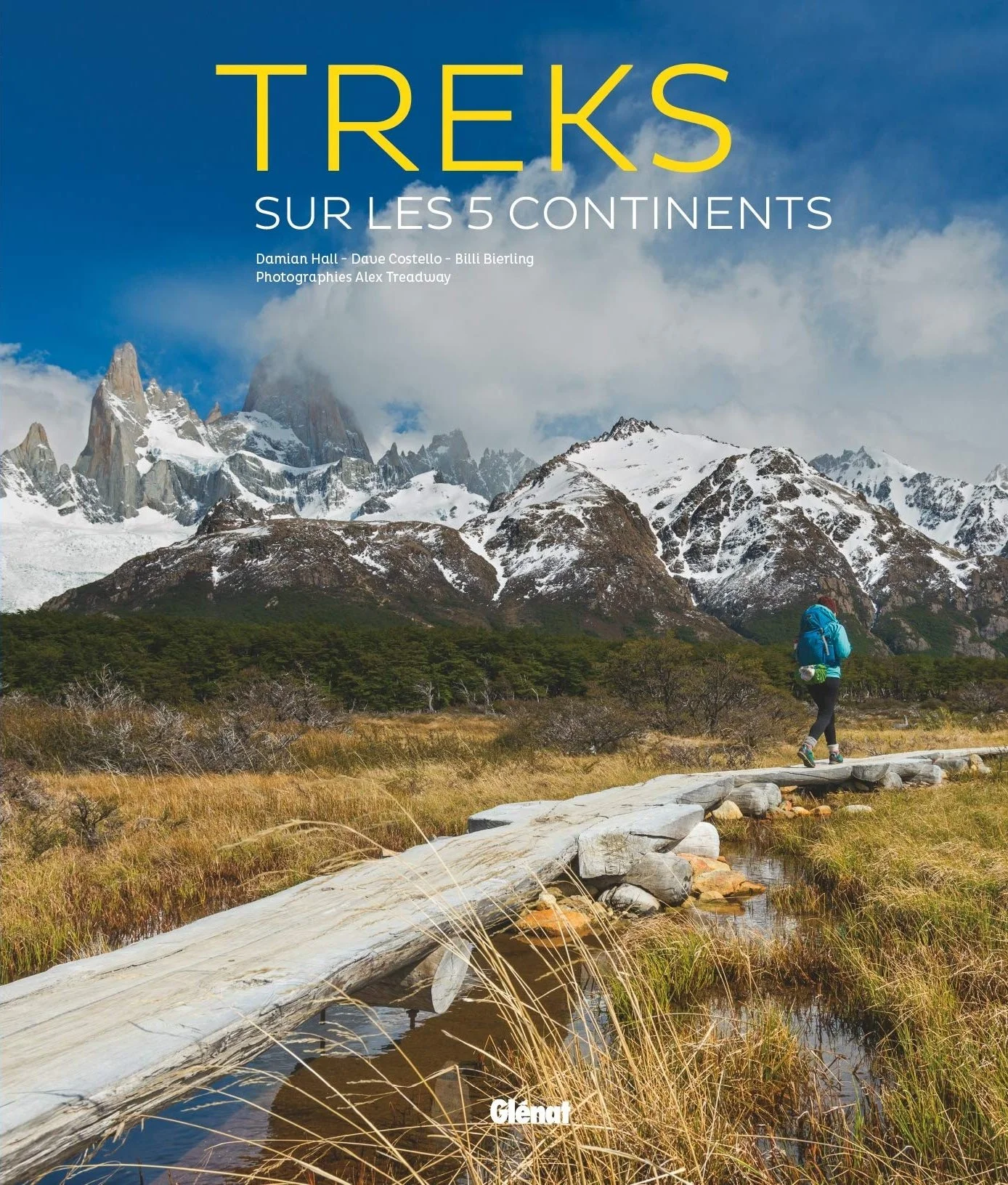 A hiker walks along a wooden trail in a mountainous landscape with snow-capped peaks and a partly cloudy sky.