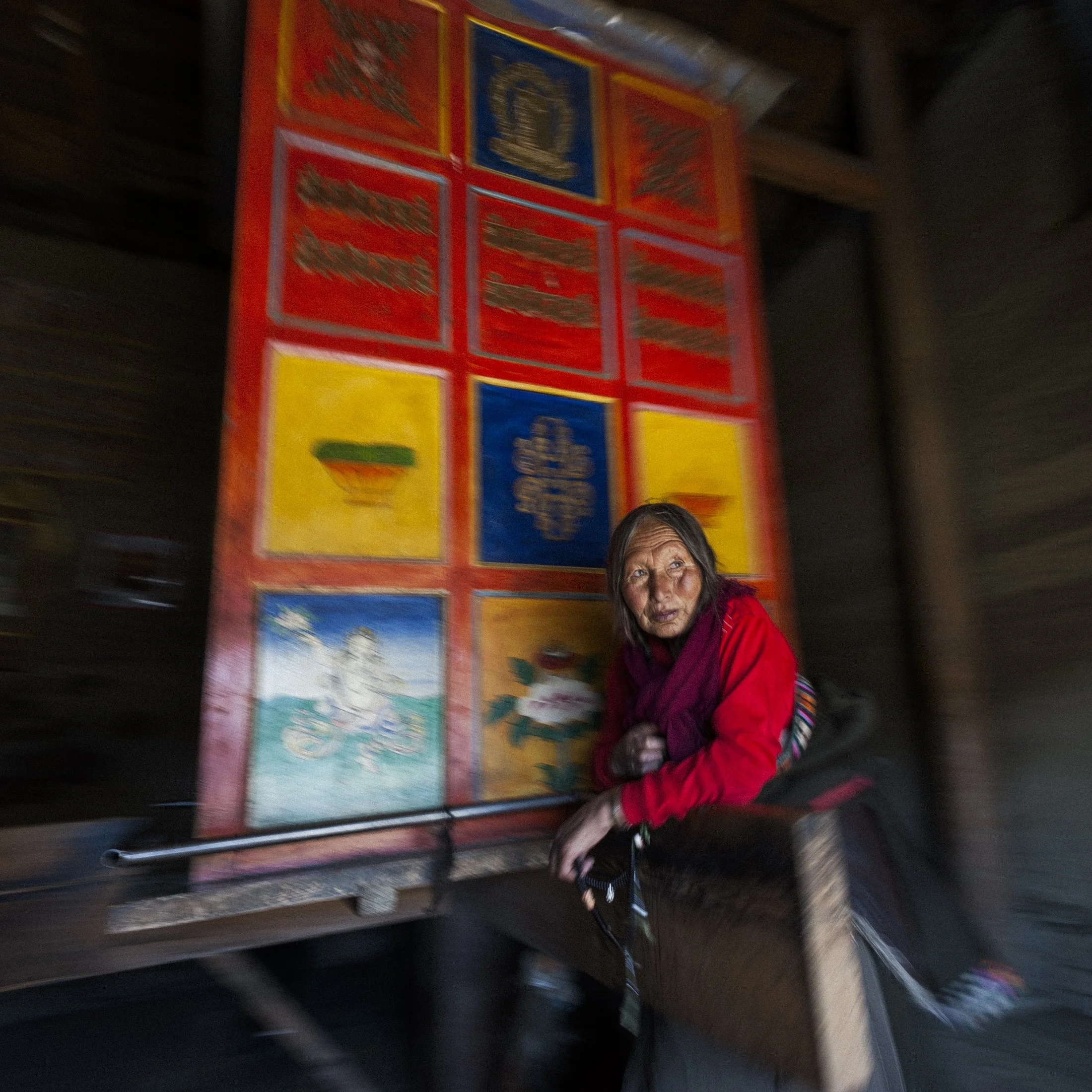 A Tibetan woman turns a large colourful prayer wheel in a remote part of China. She is wearing traditional Tibetan clothes and looking out towards the light. There is some blurred motion on the prayer wheell.