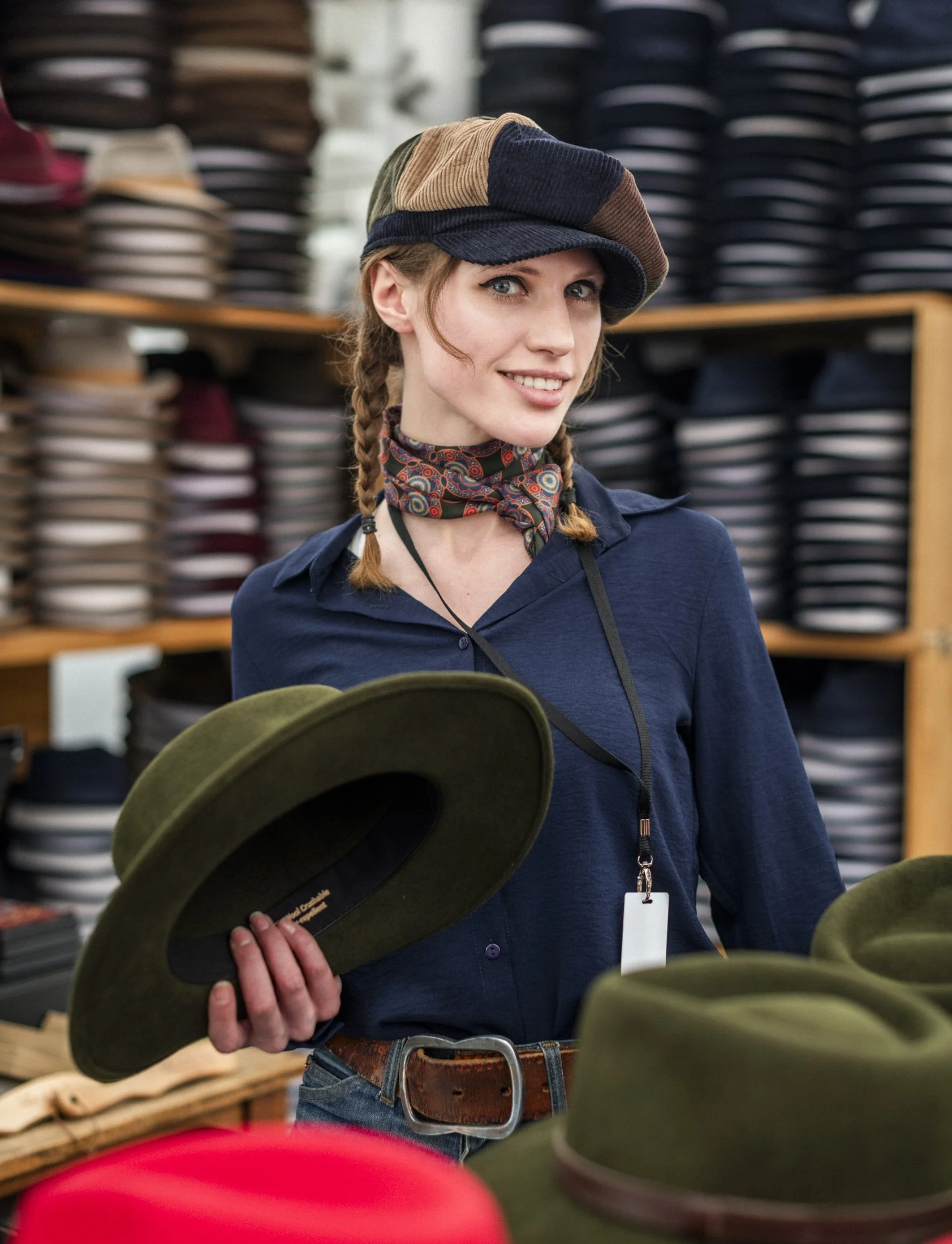 A beautiful woman working in a hat shop models one of the hats and holds another in her hand. The background is full of piles of hats on display. 