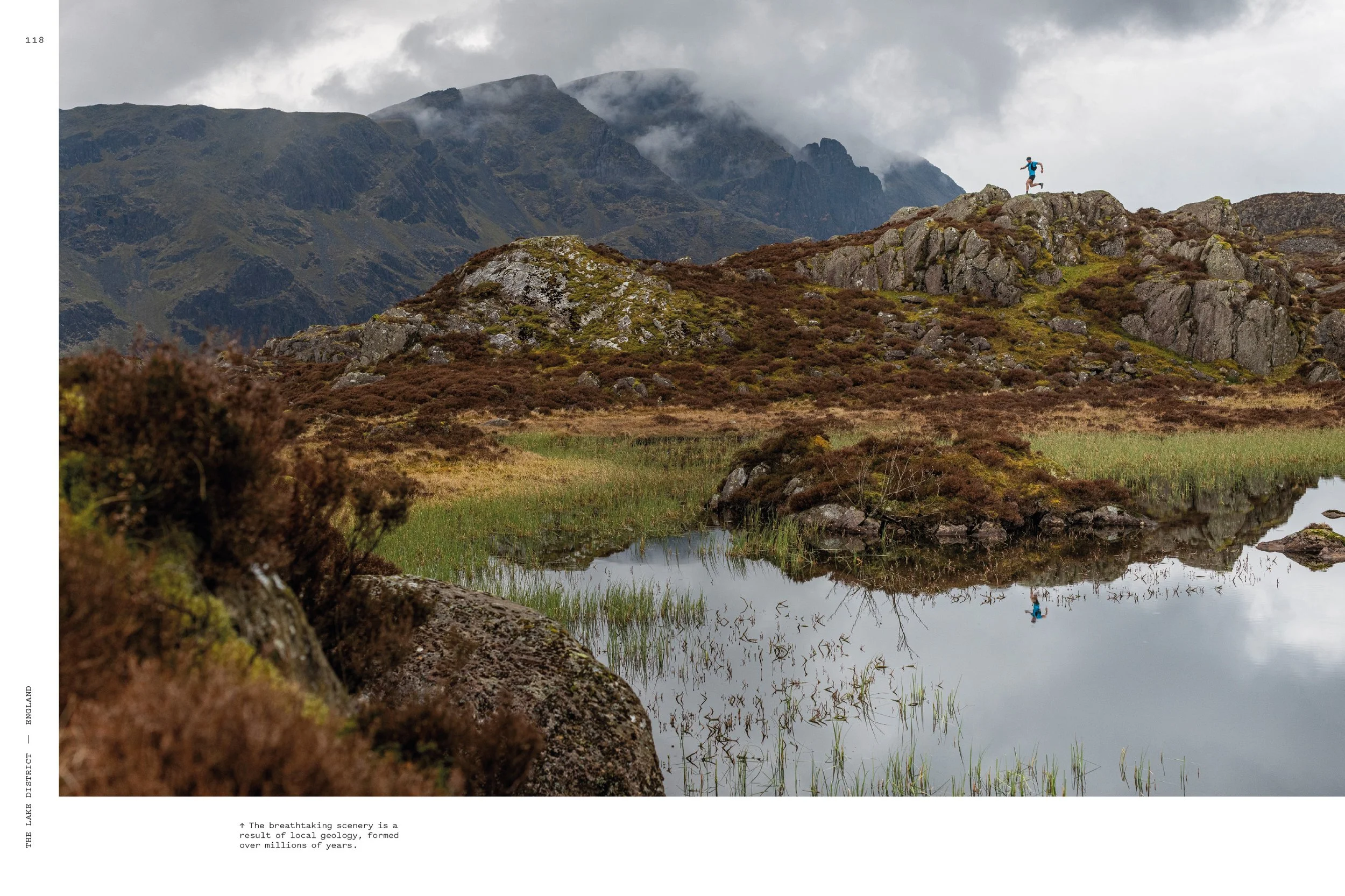 A scenic landscape of mountains with cloudy sky, a small body of water with reflected bushes, and a person in blue running along rocky terrain