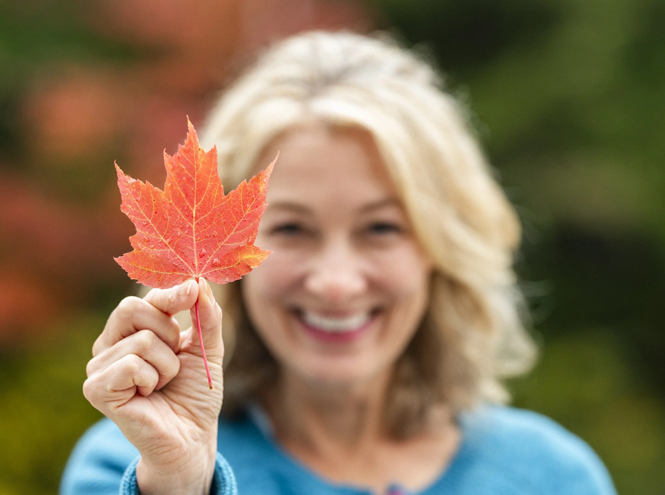 A woman holds up a Maple leaf and smiles as she shows it for the camera. The leaf has turned red indicating Autumn. It looks like she's showing the leaf to represent Canada. 