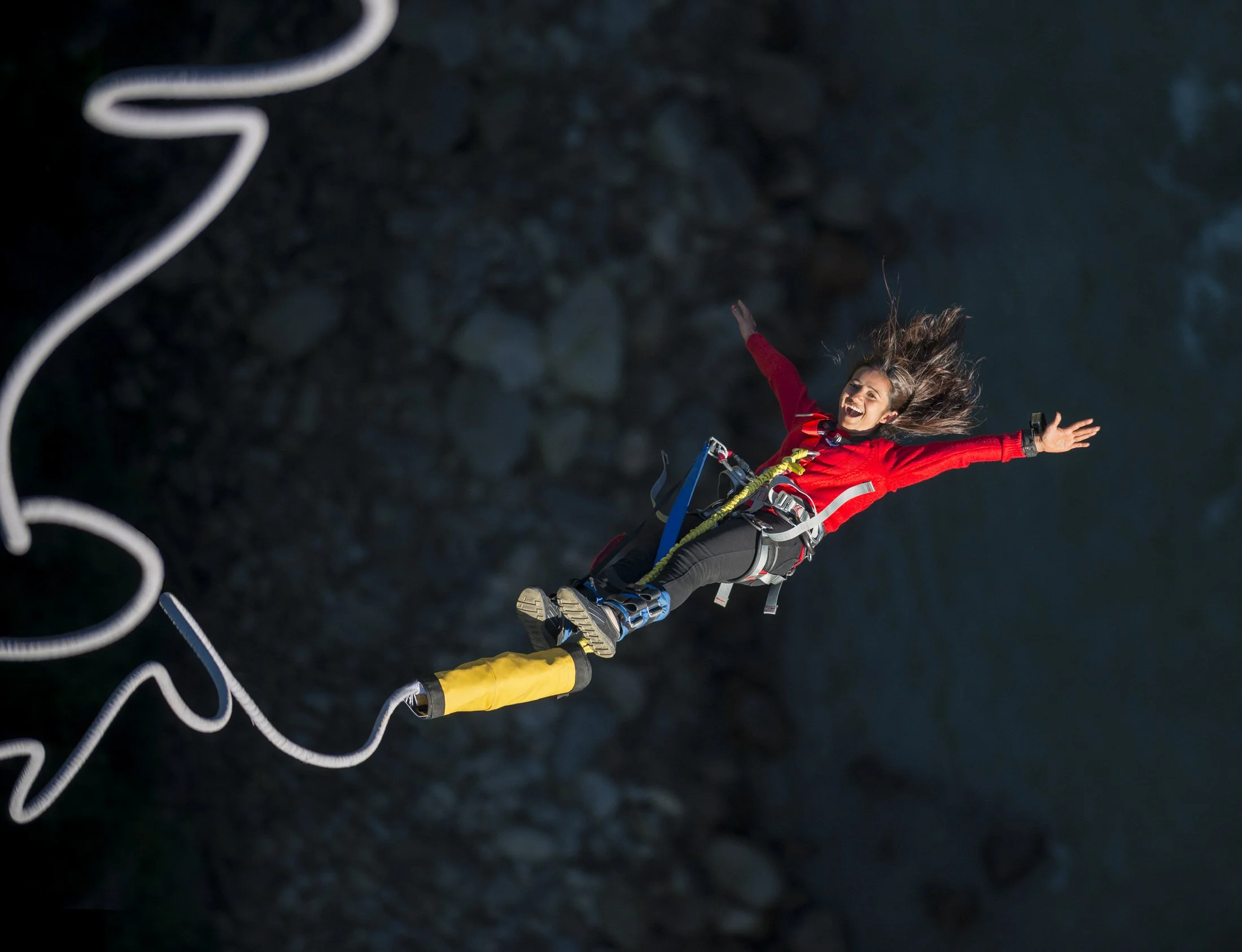 A girls screams and smiles with delight as she looks back towards up towards the bridge she has done a bungee jump from. The background is dark and the bungee rope is making a nice curving line contrasting the background. 