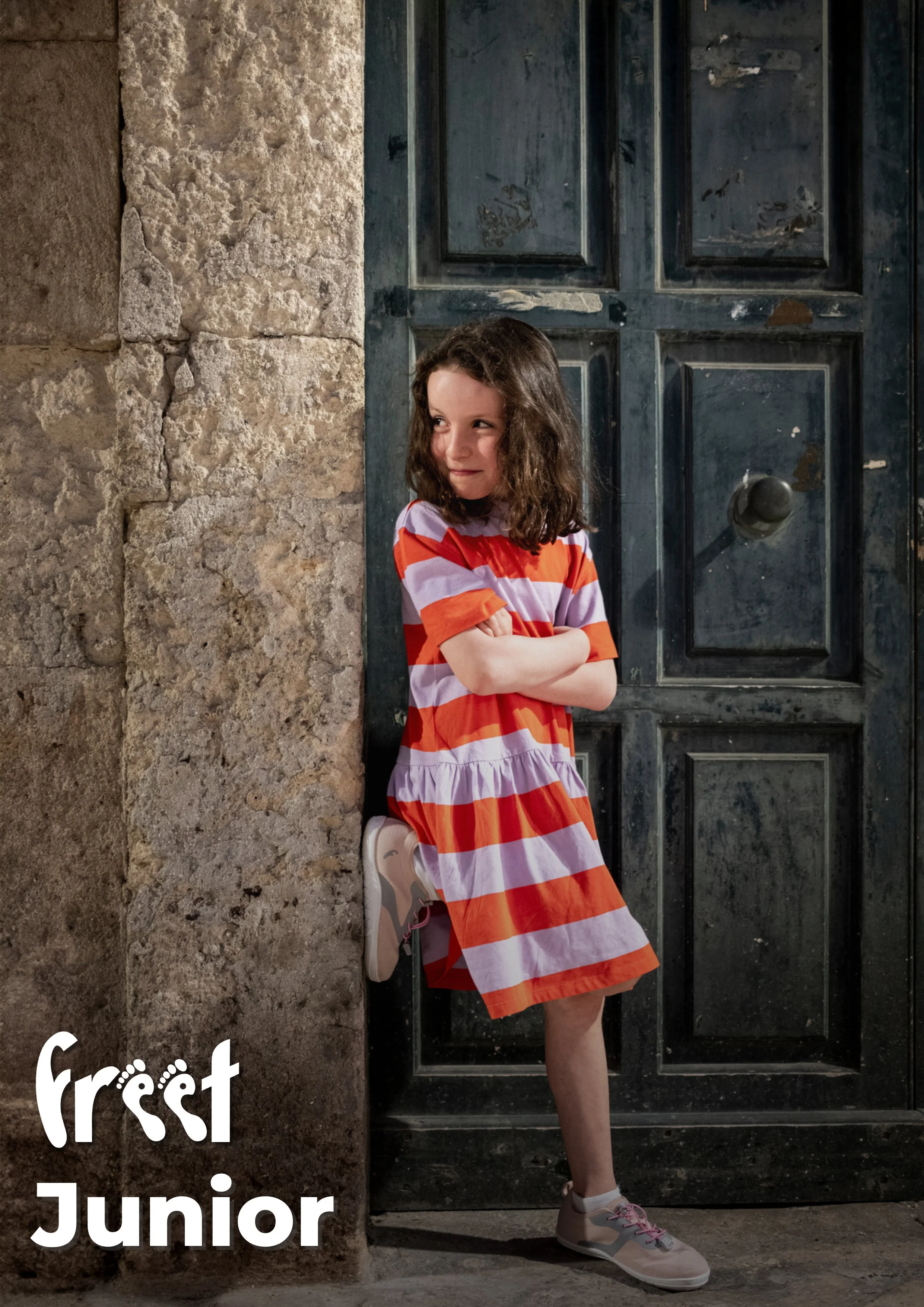 Young girl with curly hair wearing a red and white striped dress, white sneakers, standing with arms crossed and one foot against a stone wall, in front of a black door.