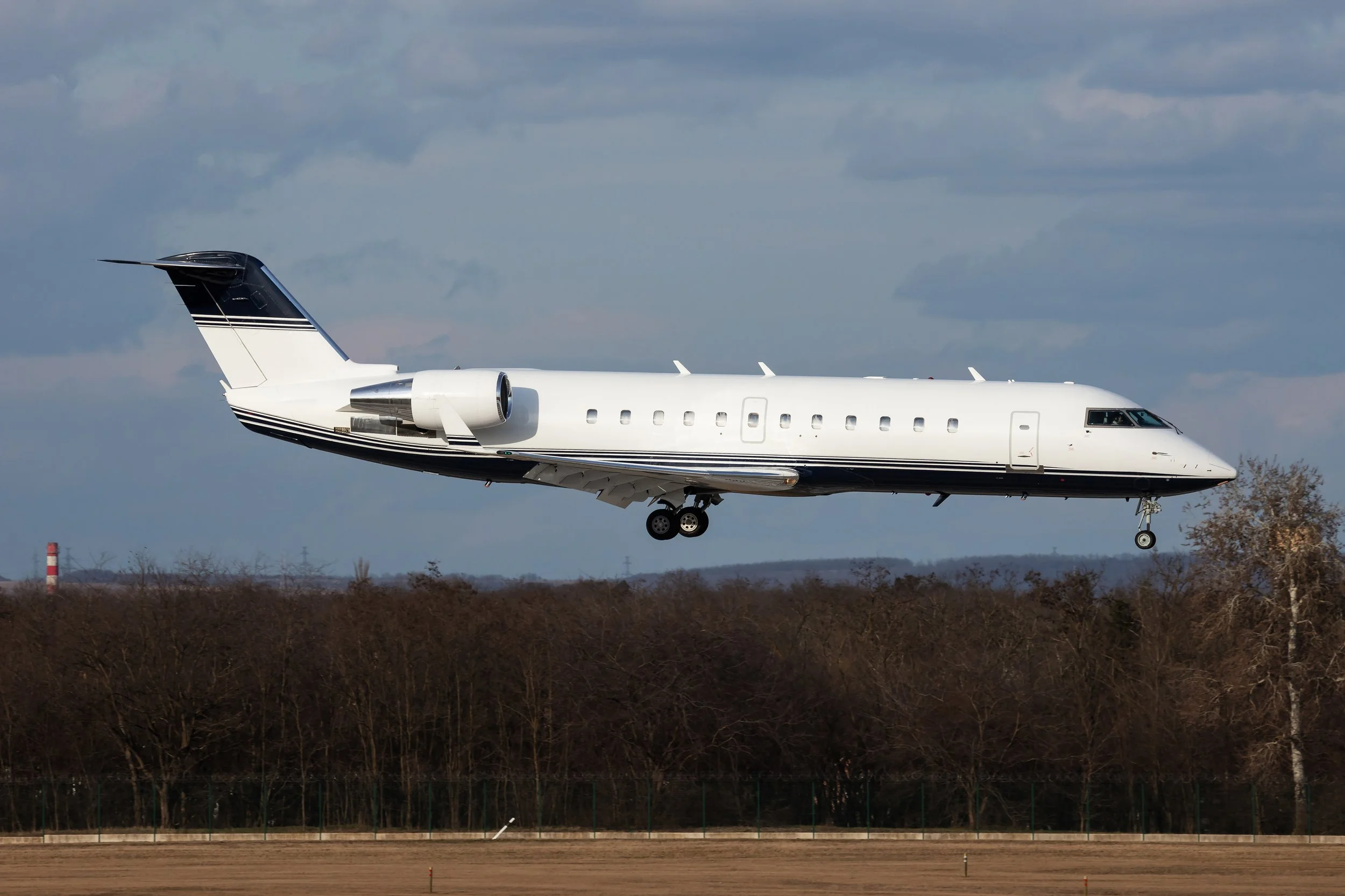A private jet airplane in mid-flight, flying low over a landscape of trees with a cloudy sky in the background.