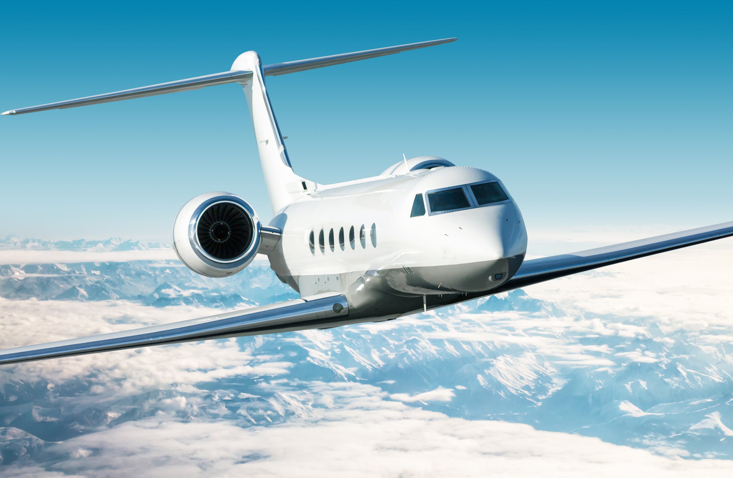 A white private jet flying high above snow-covered mountains under a blue sky.