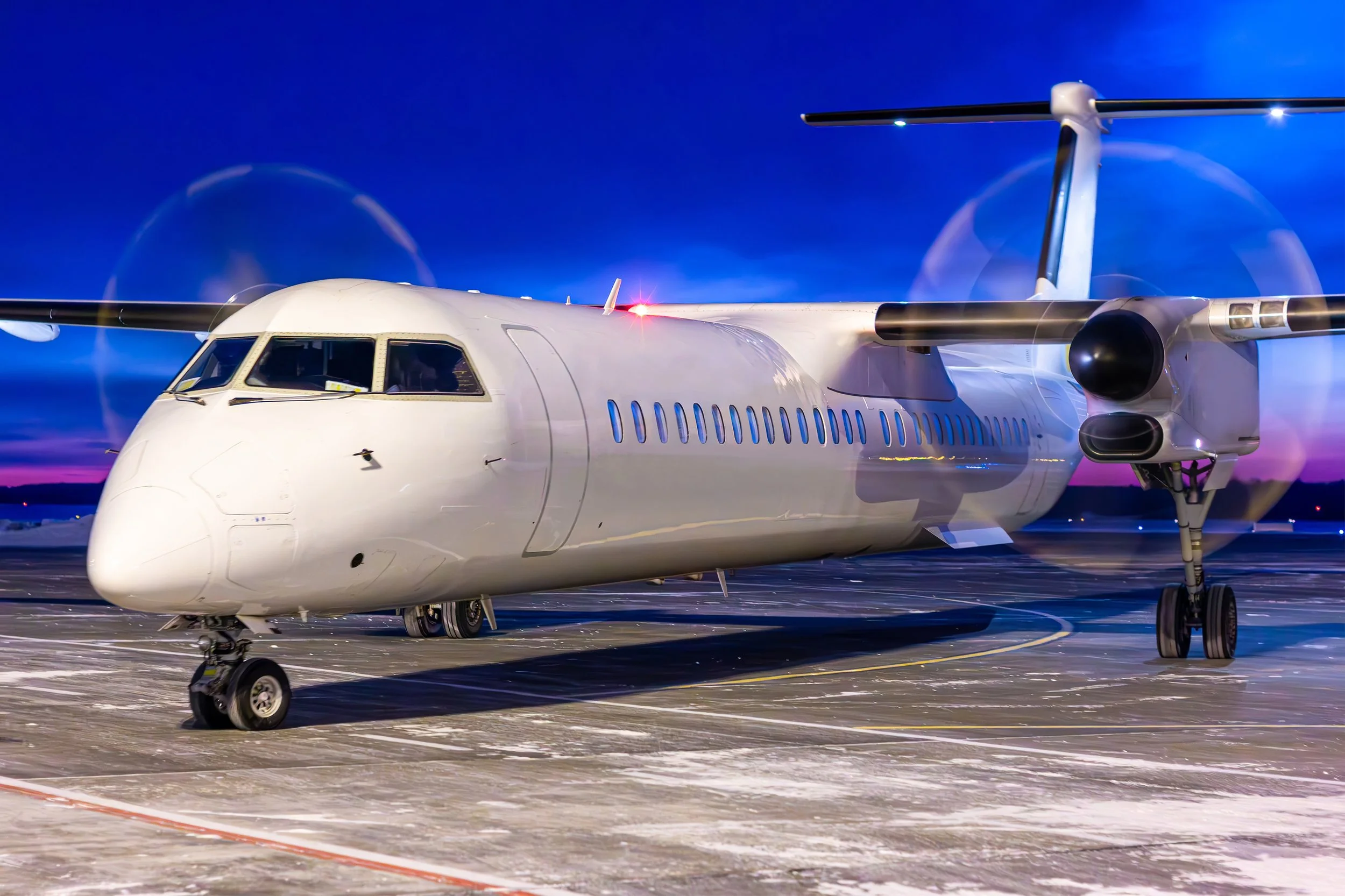 A white futuristic aircraft with propellers on a tarmac at dusk, illuminated by spotlights and glowing lights on the aircraft, against a dark blue sky.