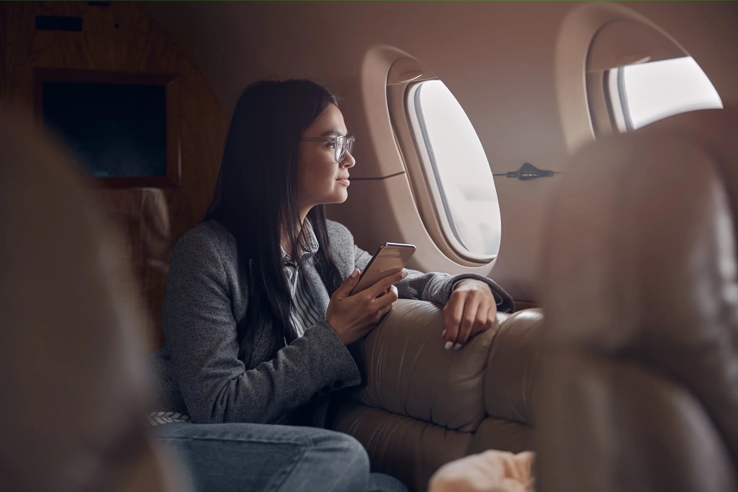 A woman with glasses sitting on an airplane, looking out the window and holding a smartphone in her hand.
