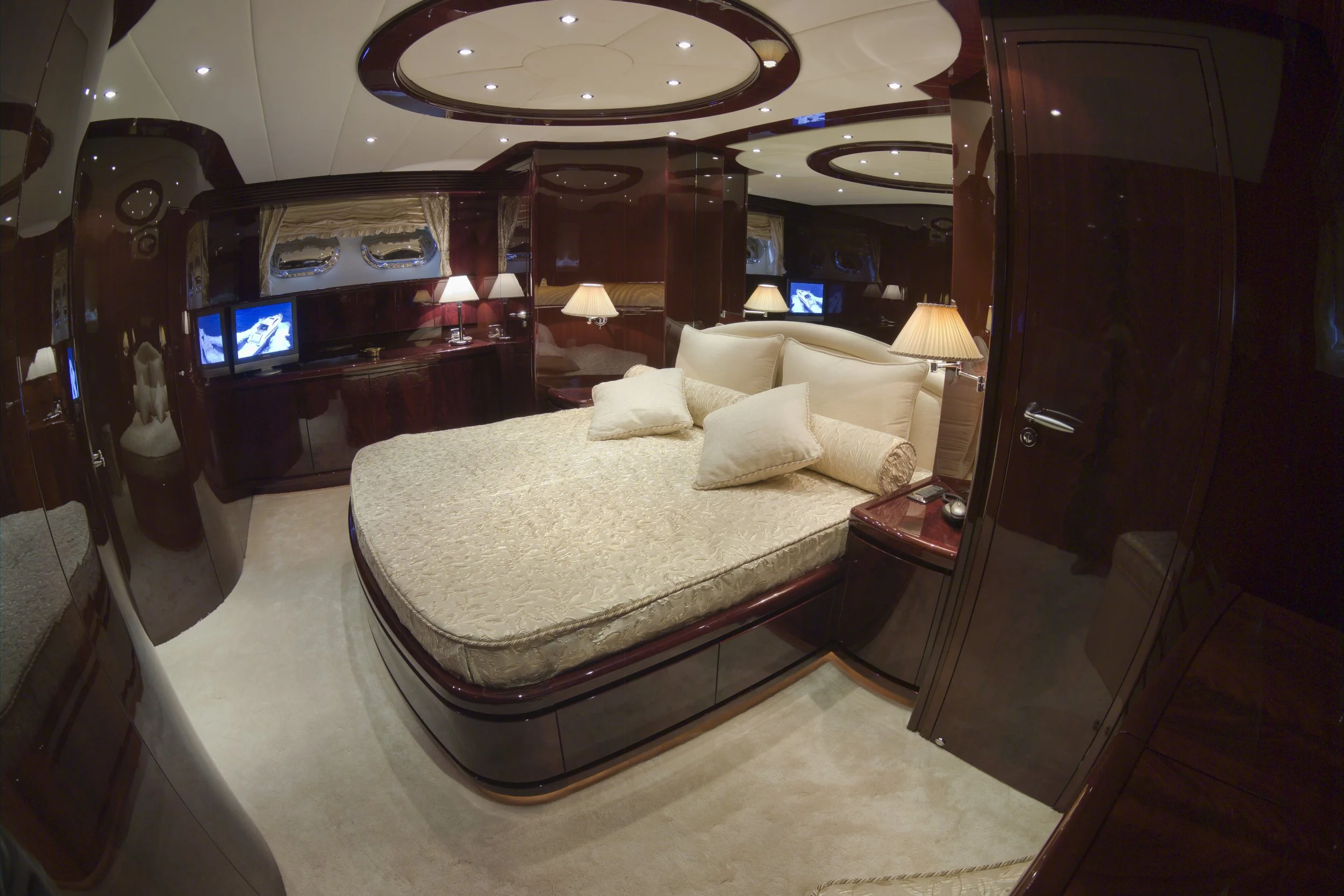 Luxurious bedroom interior with a beige bed, multiple pillows, dark wood finish cabinets, two table lamps, and a TV mounted on the wall.