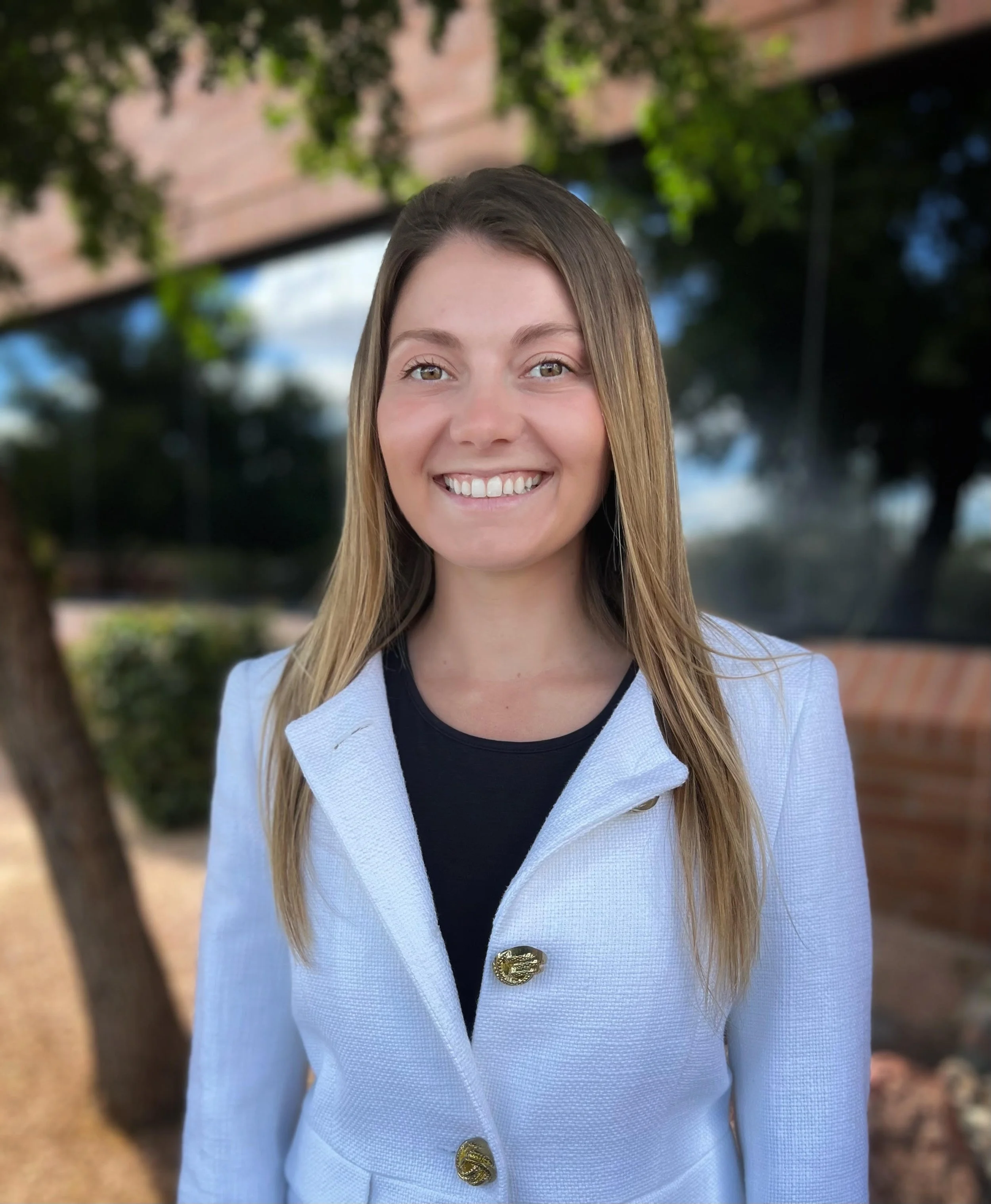 A young woman with long blonde hair smiling outdoors, wearing a light-colored blazer with gold buttons and a black shirt underneath.