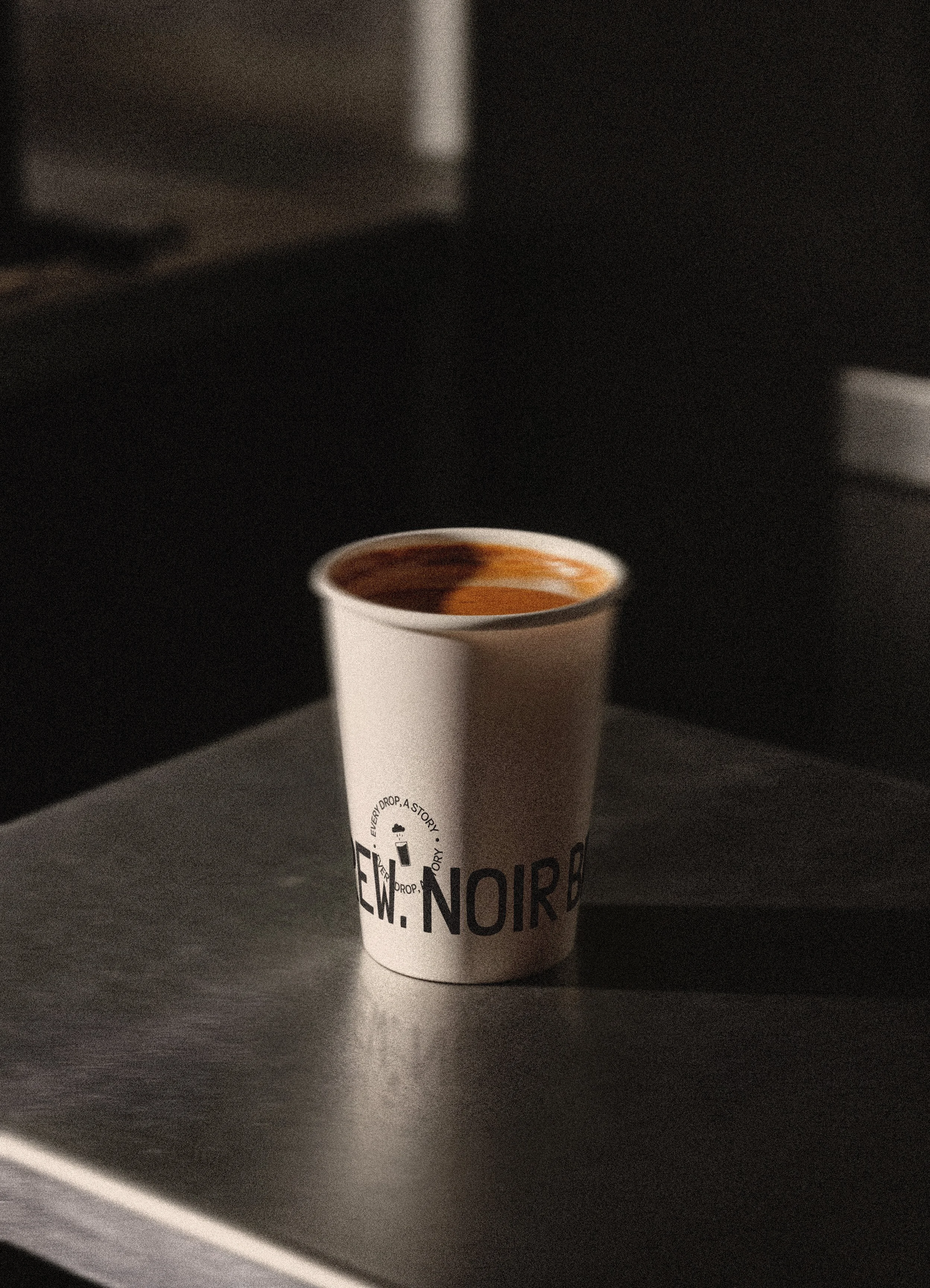 Mise en situation de l'identité visuelle réalisée pour le coffee shop Noir Brew. Le logo principal est aposé sur un verre en papier blanc rempli de café noir sur un plan de travail en métal chromé, avec un fond sombre.